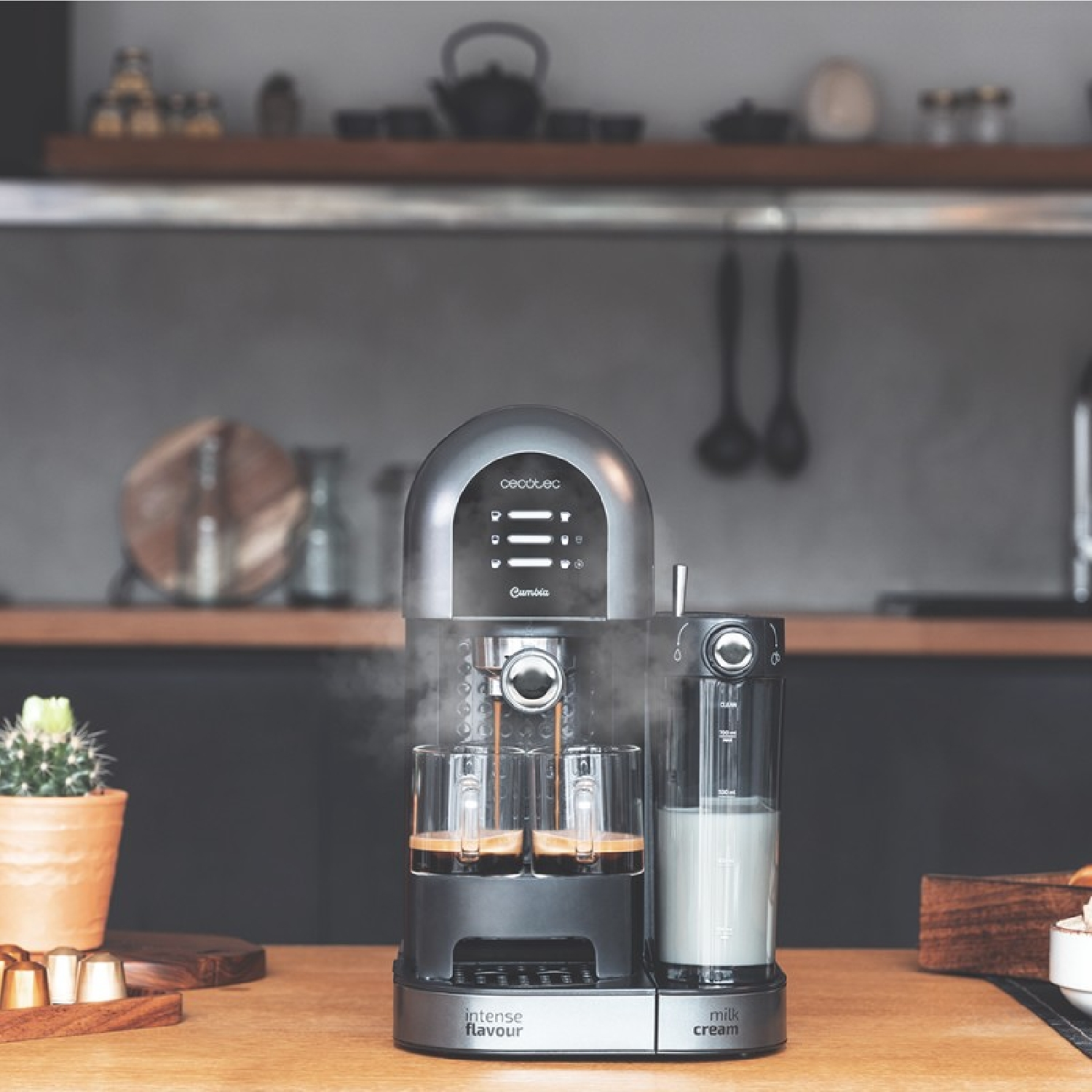 Una macchina da caffè con due espressi e montalatte su un tavolo di legno. Un cactus e capsule sono visibili a sinistra.