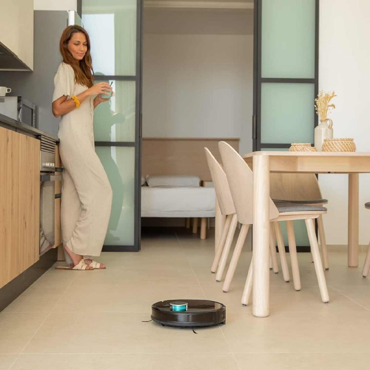 Frau in der Küche mit Getränk, Roboter saugt. Tisch und Stühle im Hintergrund. Schlafzimmer durch Schiebetür sichtbar.