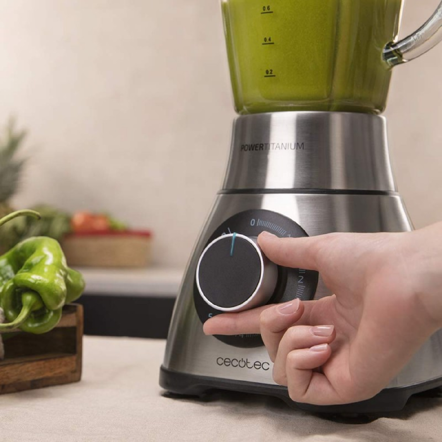 Una mano gira el dial de una licuadora llena de líquido verde. Pimientos verdes y verduras en el fondo.