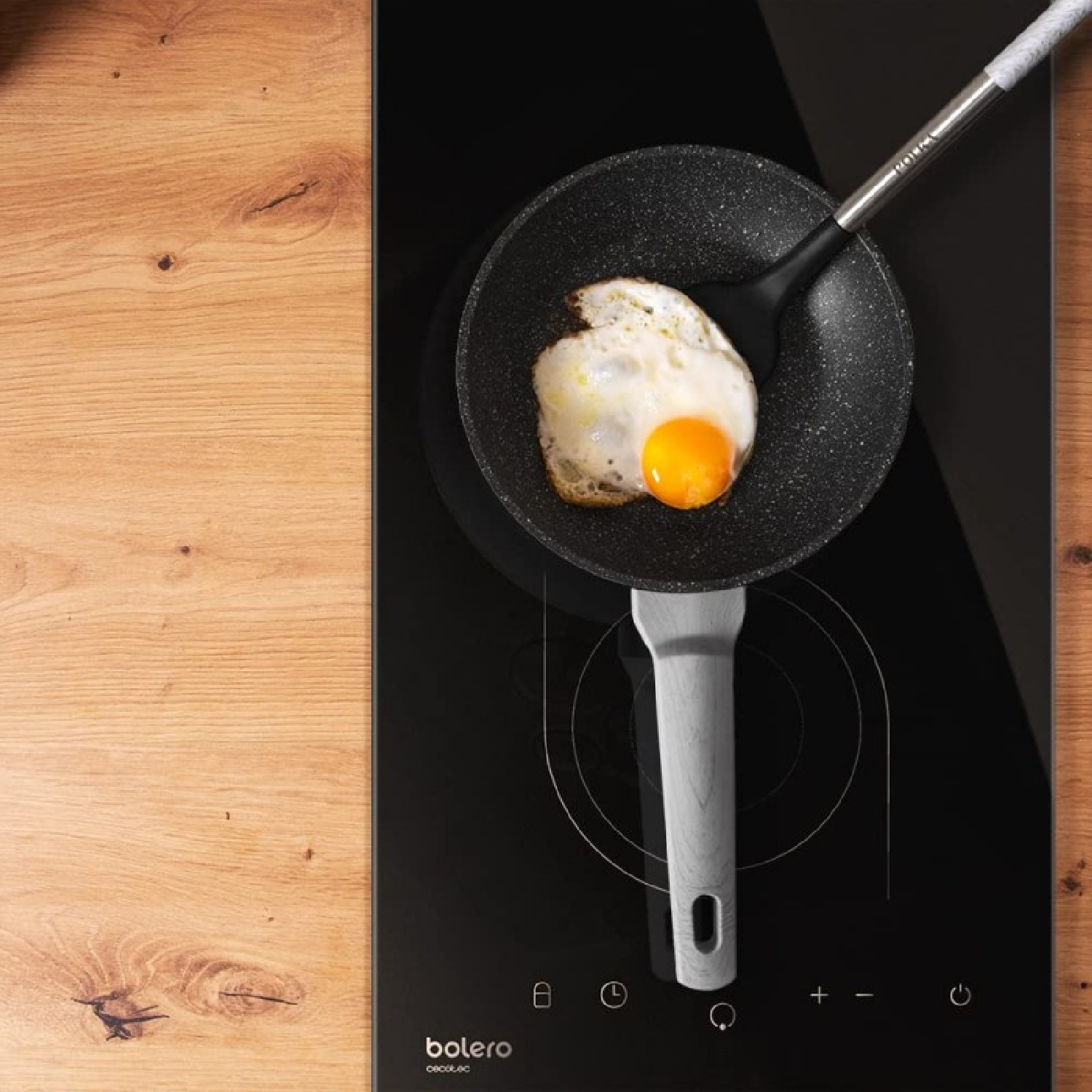 A fried egg in a pan on a black stove. A spatula is lifting the egg. Wooden table on the side.