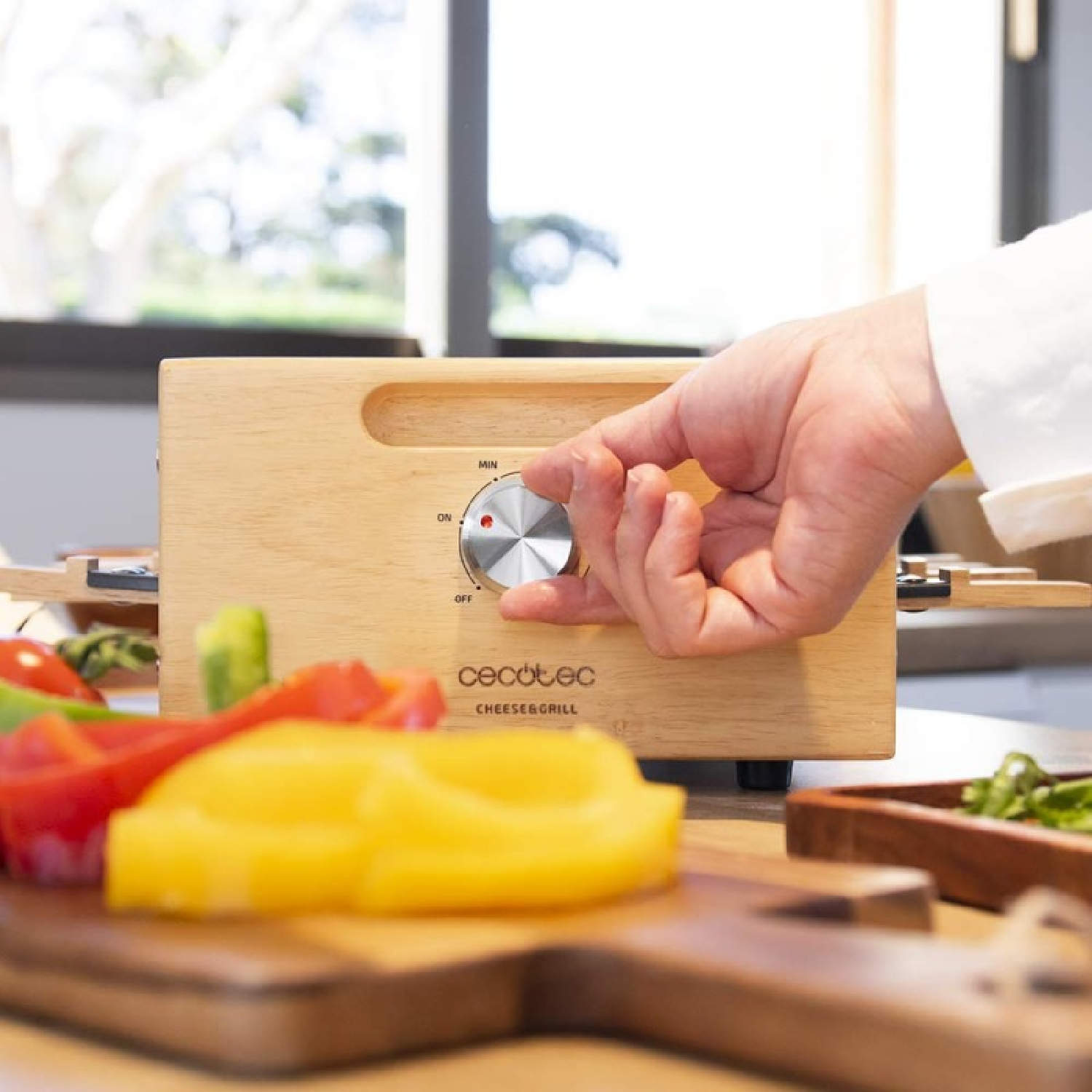 Ręka przekręca pokrętło drewnianego grilla do sera. Pokrojona papryka i drewniana taca z zielenią na pierwszym planie.