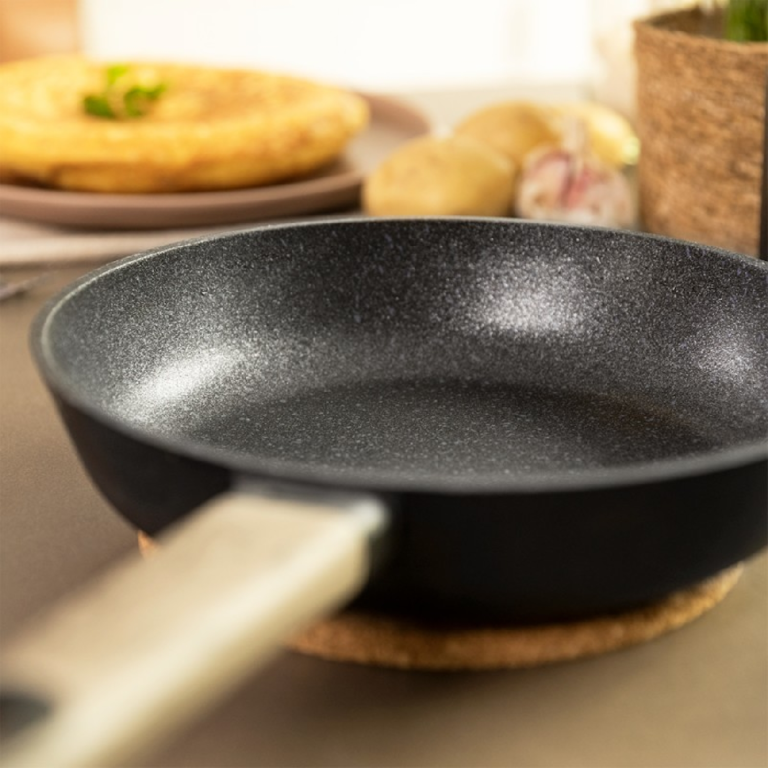 Primo piano di una padella, con tortilla e patate sullo sfondo. La padella è su una base di sughero.