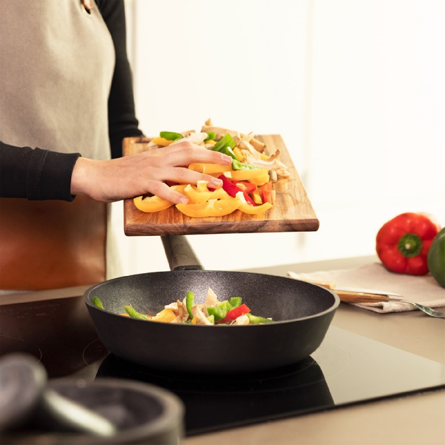 Una persona aggiunge peperoni tritati a una padella. Un tagliere di legno contiene peperoni colorati. Un peperone rosso è sul bancone.