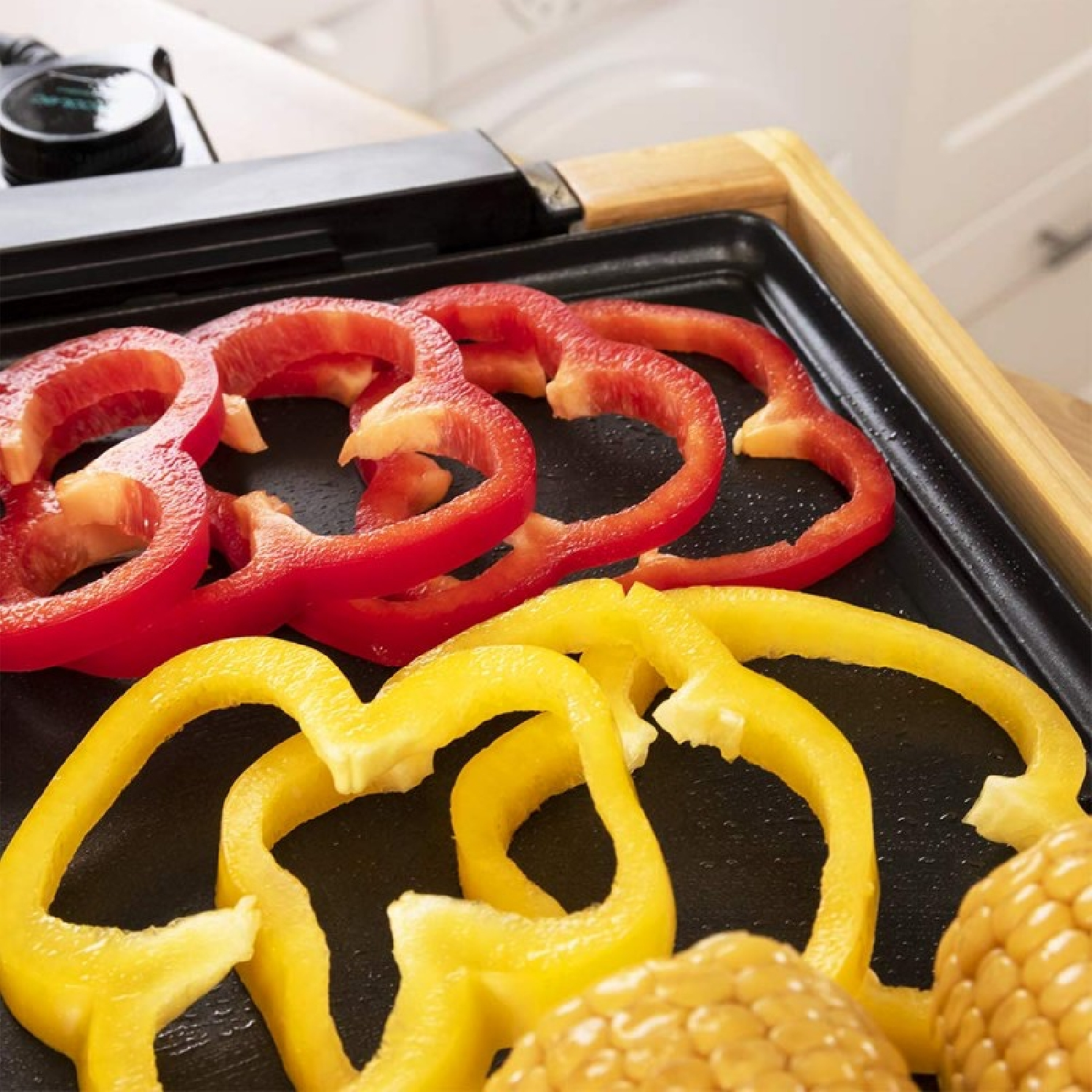 Plastry czerwonej i żółtej papryki na czarnym grillu. Widoczna kukurydza. Grill ma drewnianą ramę i pokrętło sterowania.
