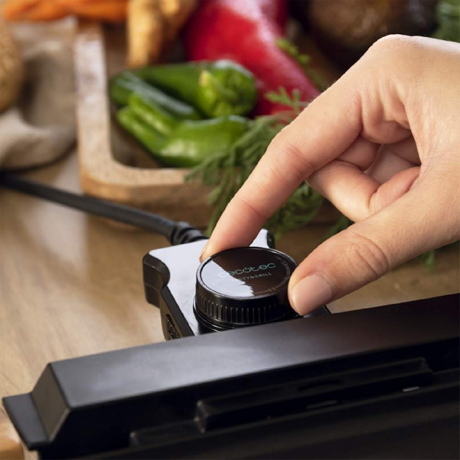 Ręka reguluje czarne pokrętło na grillu. Warzywa w drewnianej tacy w tle.