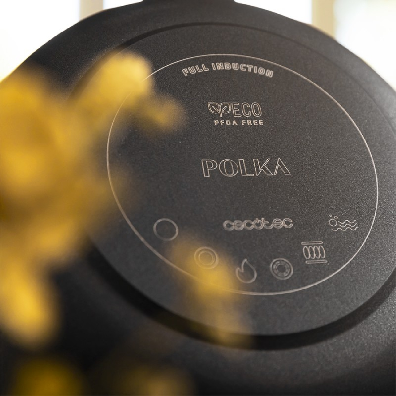 Primo piano di una padella a induzione nera. Caratteristiche: marchio 'POLKA', 'PECO PFOA FREE' e vari simboli di cottura. Sfocatura gialla in primo piano.