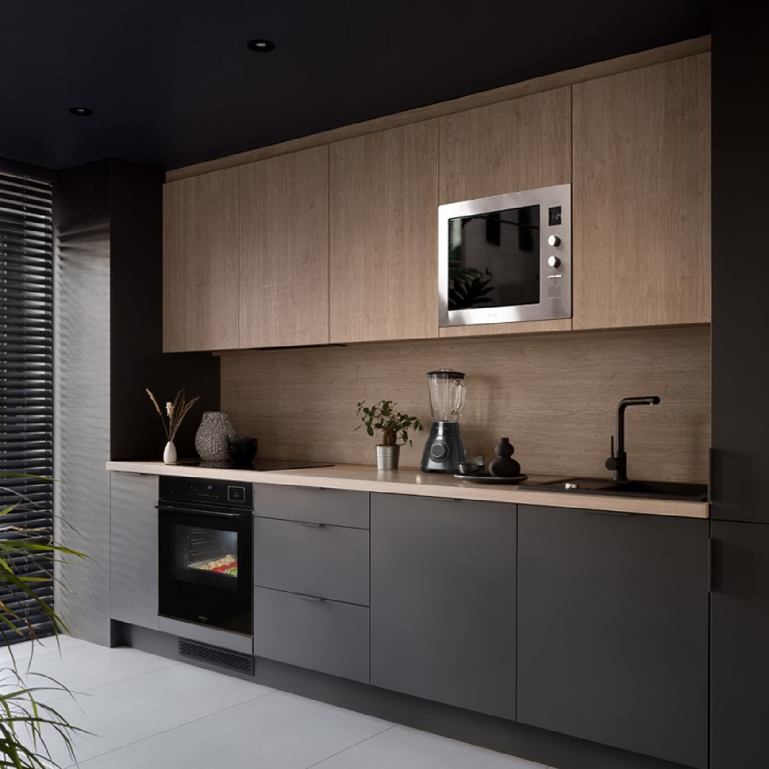 Nowoczesna kuchnia z drewnianymi szafkami, kuchenką mikrofalową, piekarnikiem i blenderem. Ciemnoszare szafki dolne i czarny zlew. Widoczne rośliny i dekoracje.