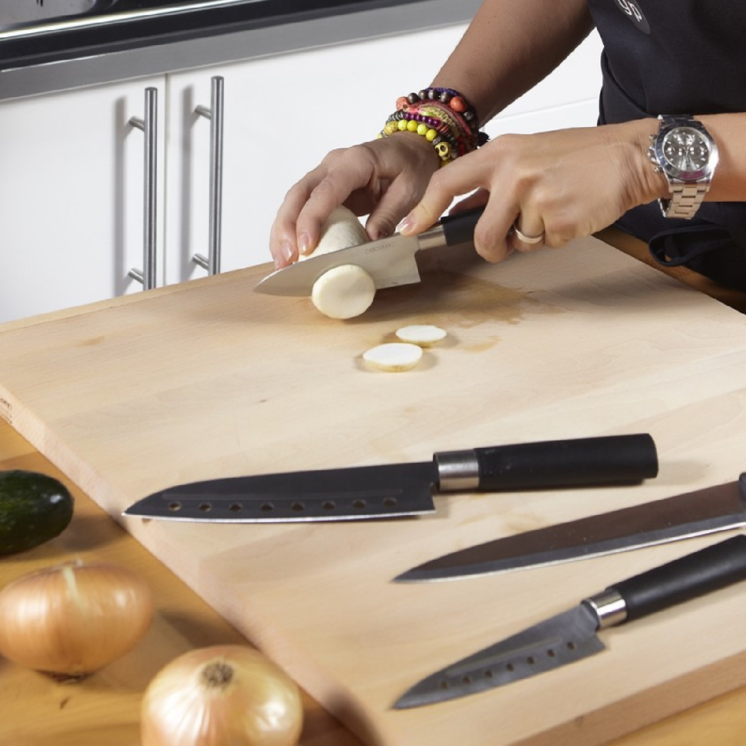 Osoba kroi cebulę na drewnianej desce. Kilka noży i inne cebule znajdują się na pierwszym planie.