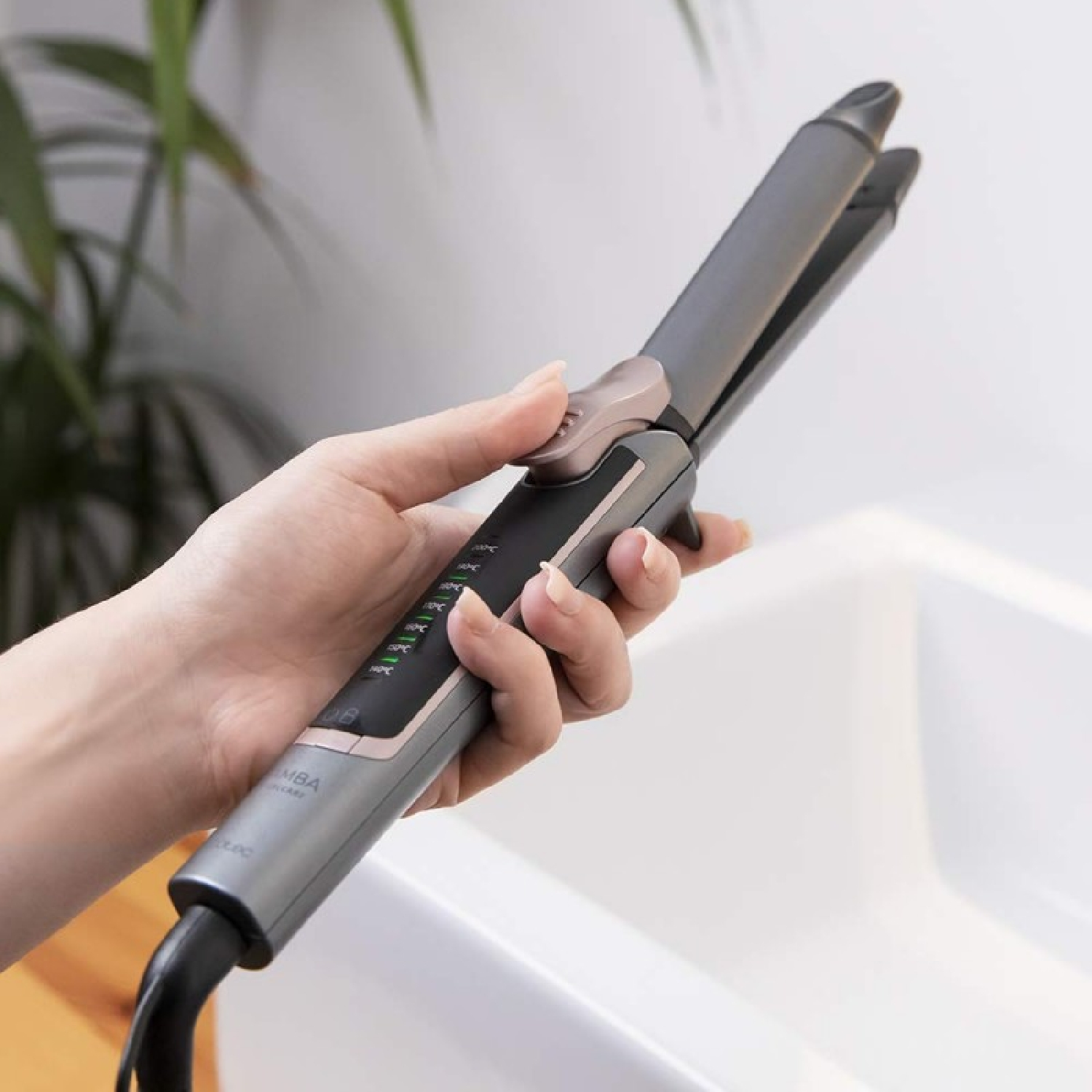 Una mano sostiene una plancha de pelo gris y negro. El fondo tiene un lavabo blanco y una planta verde borrosa.