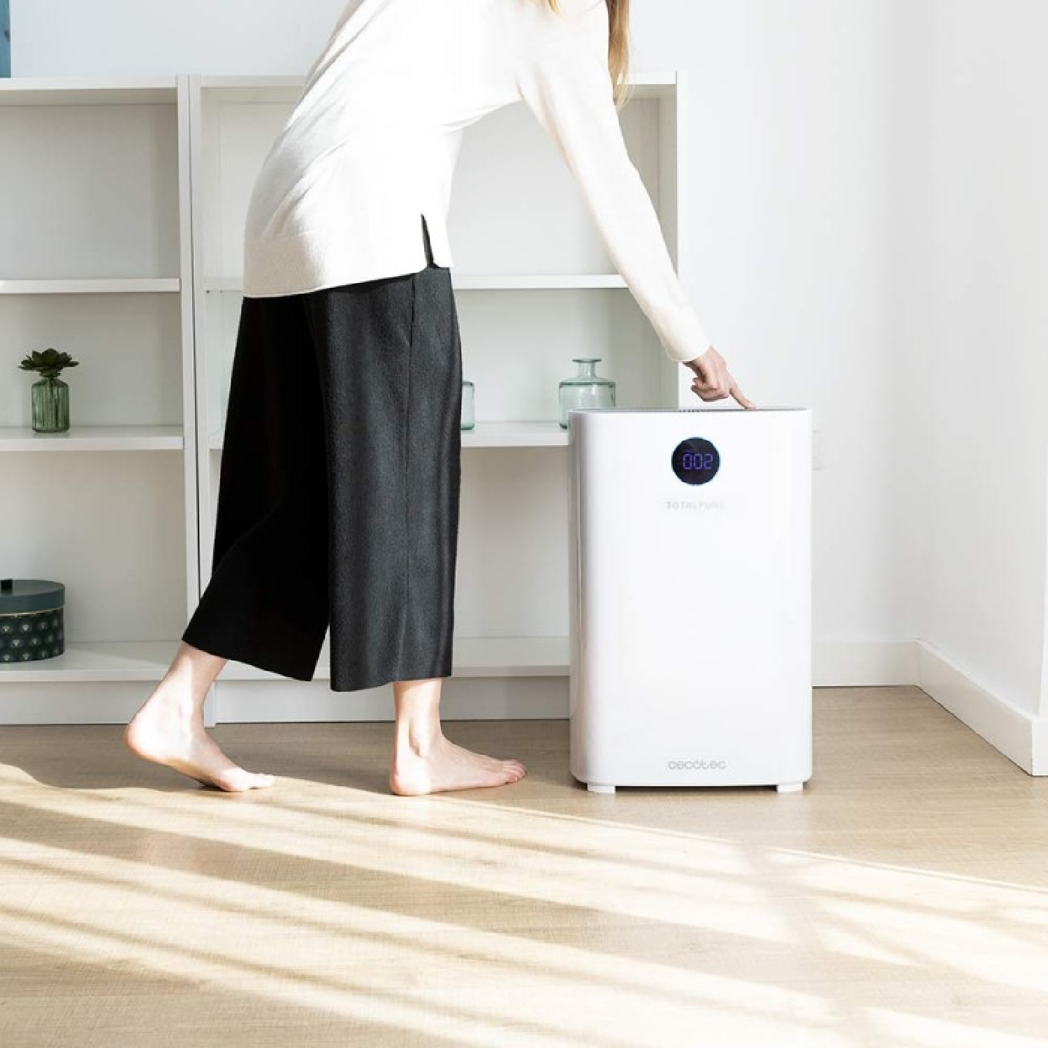 Una donna con pantaloni neri e camicia bianca tocca un purificatore d'aria bianco. Scaffali con piante sullo sfondo.