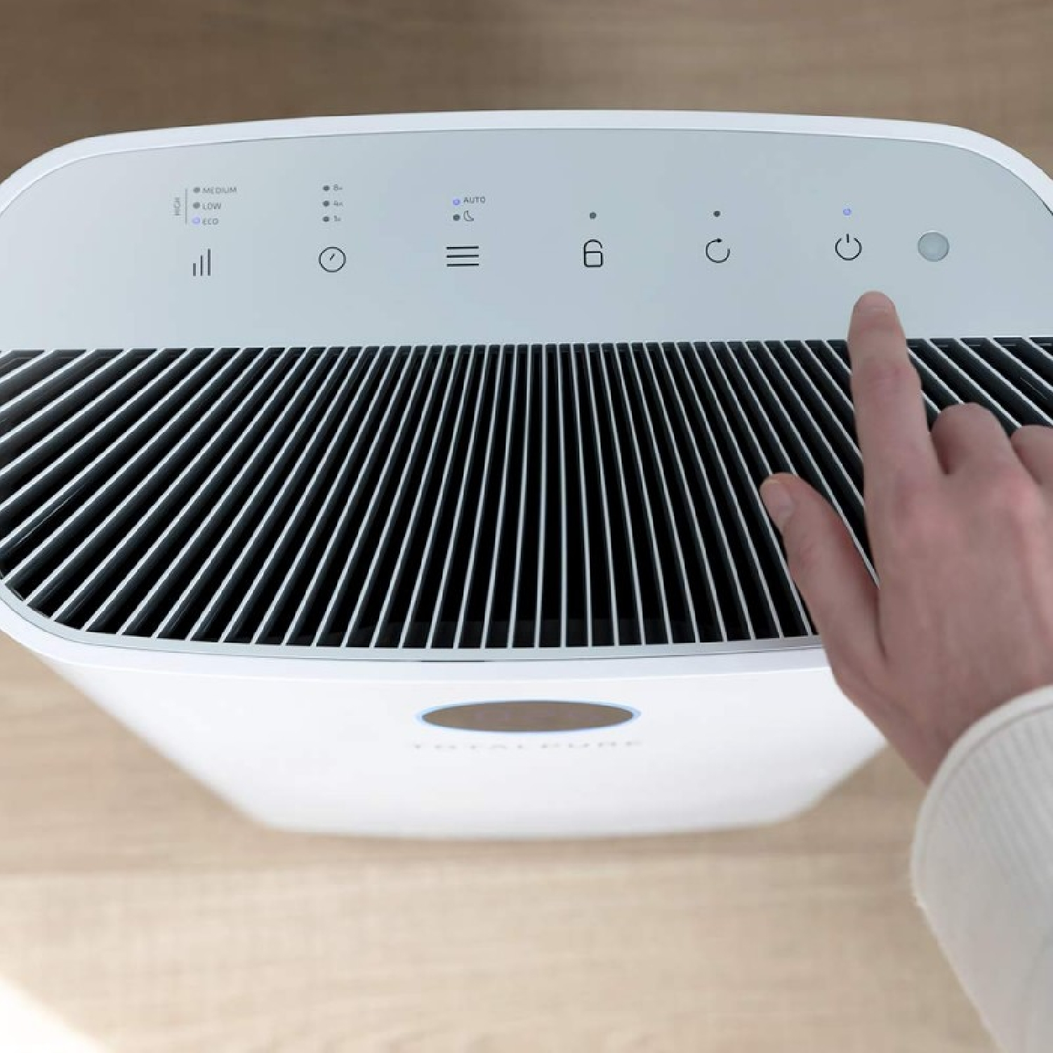 Una mano preme il pulsante di accensione di un purificatore d'aria bianco. Il purificatore è su una superficie di legno. La parte superiore ha icone di controllo.