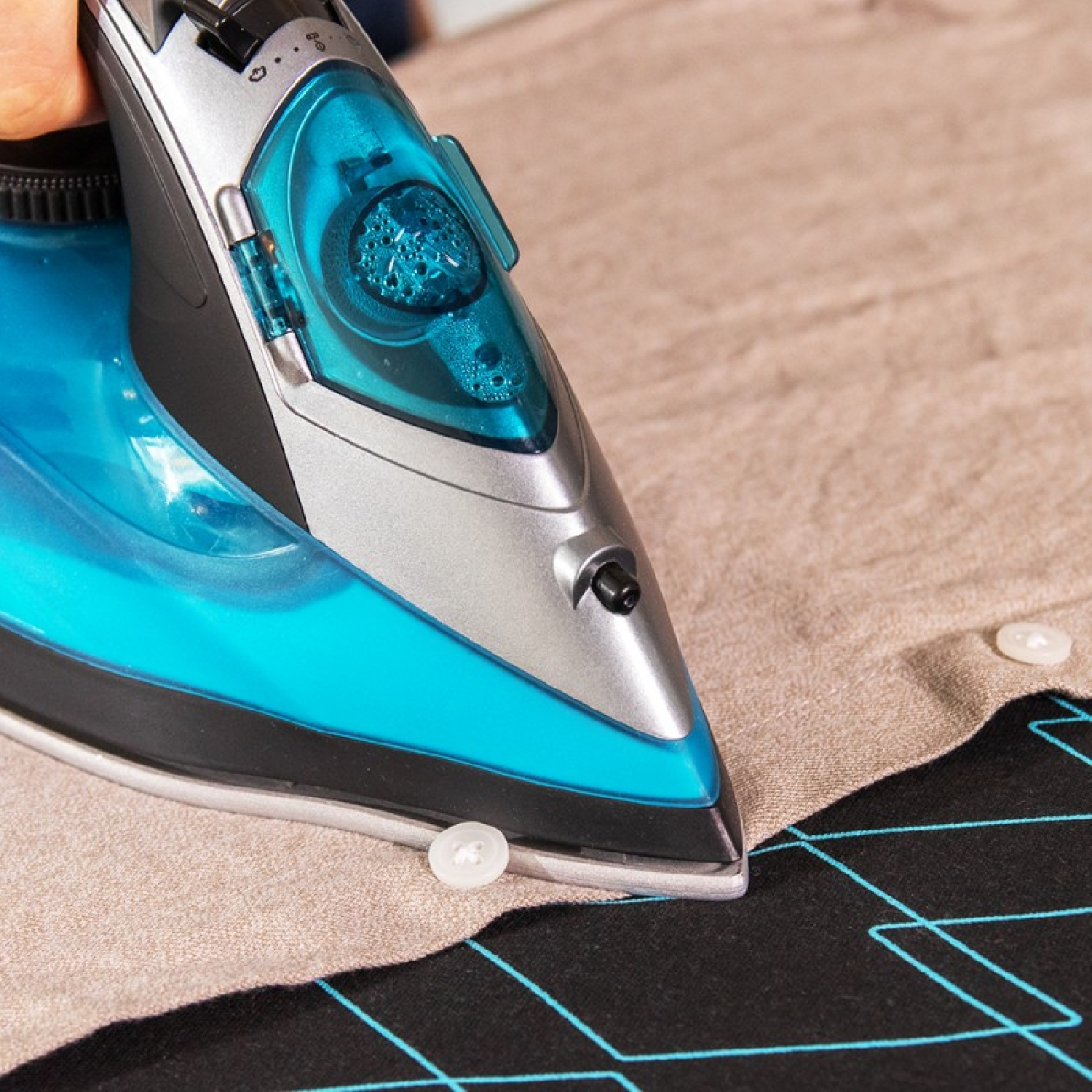 Niebiesko-srebrne żelazko prasuje beżową koszulę z białymi guzikami. Koszula leży na czarnej tkaninie z niebieskimi liniami.