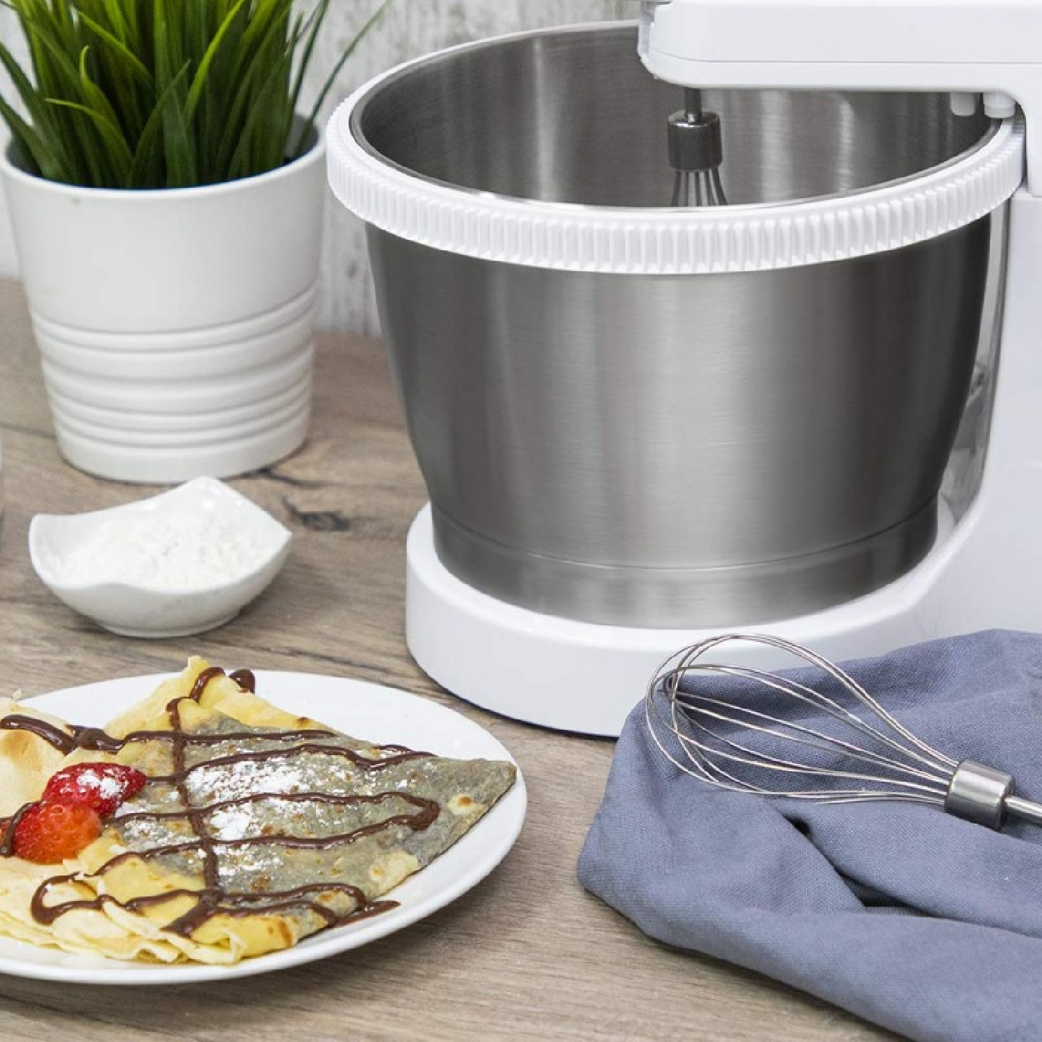 Ein Crêpe mit Erdbeeren und Schokolade, ein Mixer, eine Schüssel mit Mehl und eine Pflanze in einem weißen Topf stehen auf einem Holztisch.