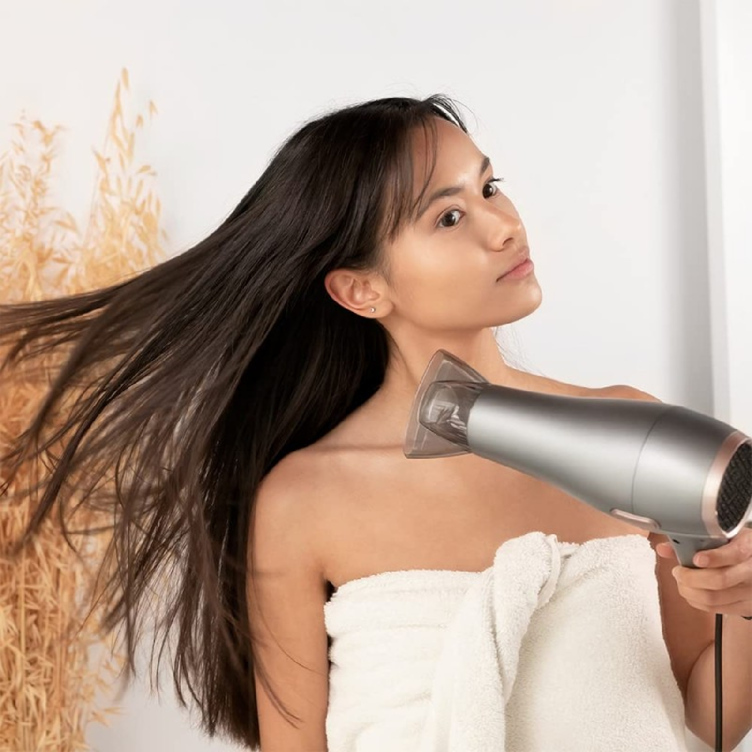 Una donna con i capelli scuri si asciuga i capelli con un asciugacapelli argentato. È avvolta in un asciugamano bianco. Lo sfondo ha piante secche.