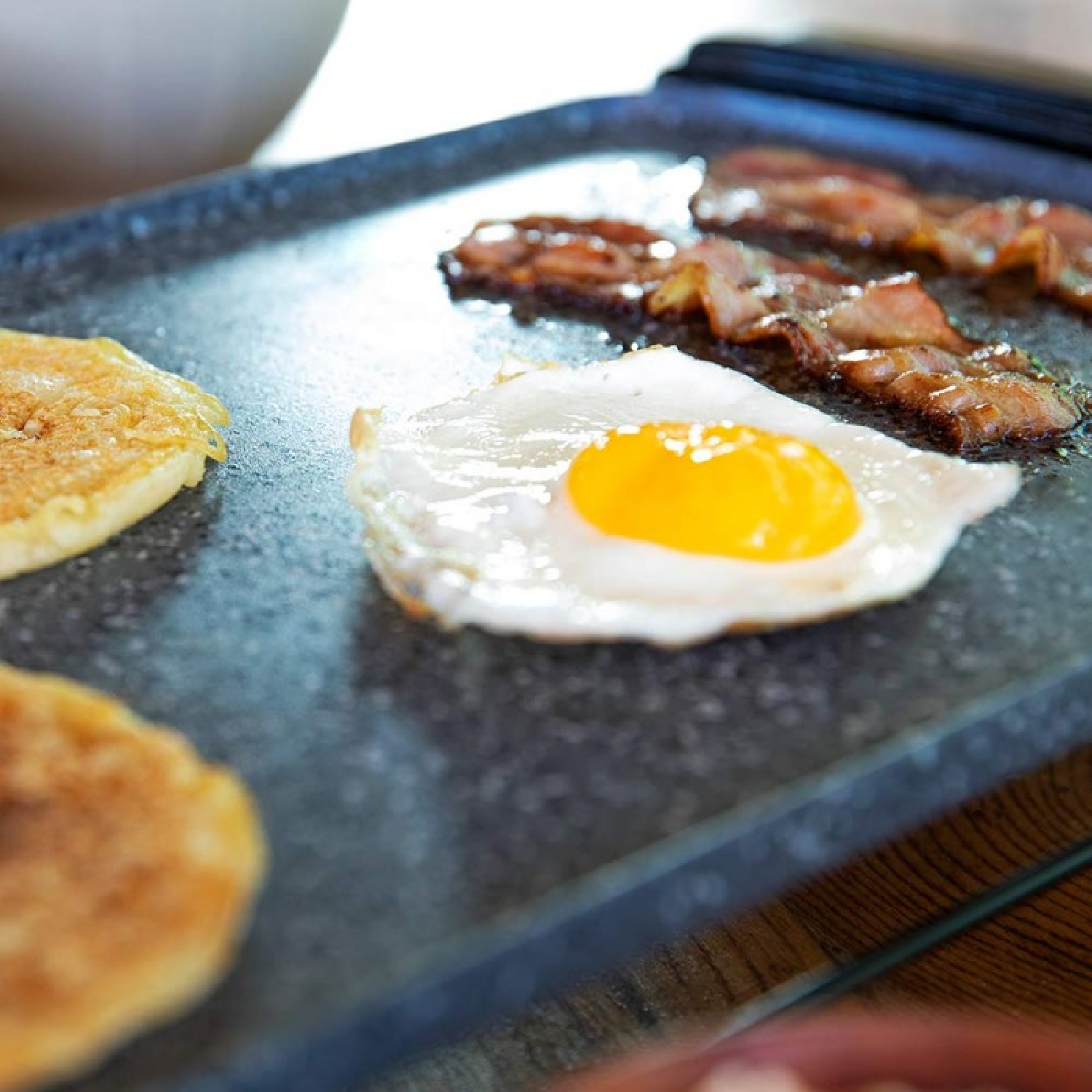 Primer plano de una plancha con tocino cocido, un huevo frito con yema amarilla y panqueques. La superficie es gris oscuro.