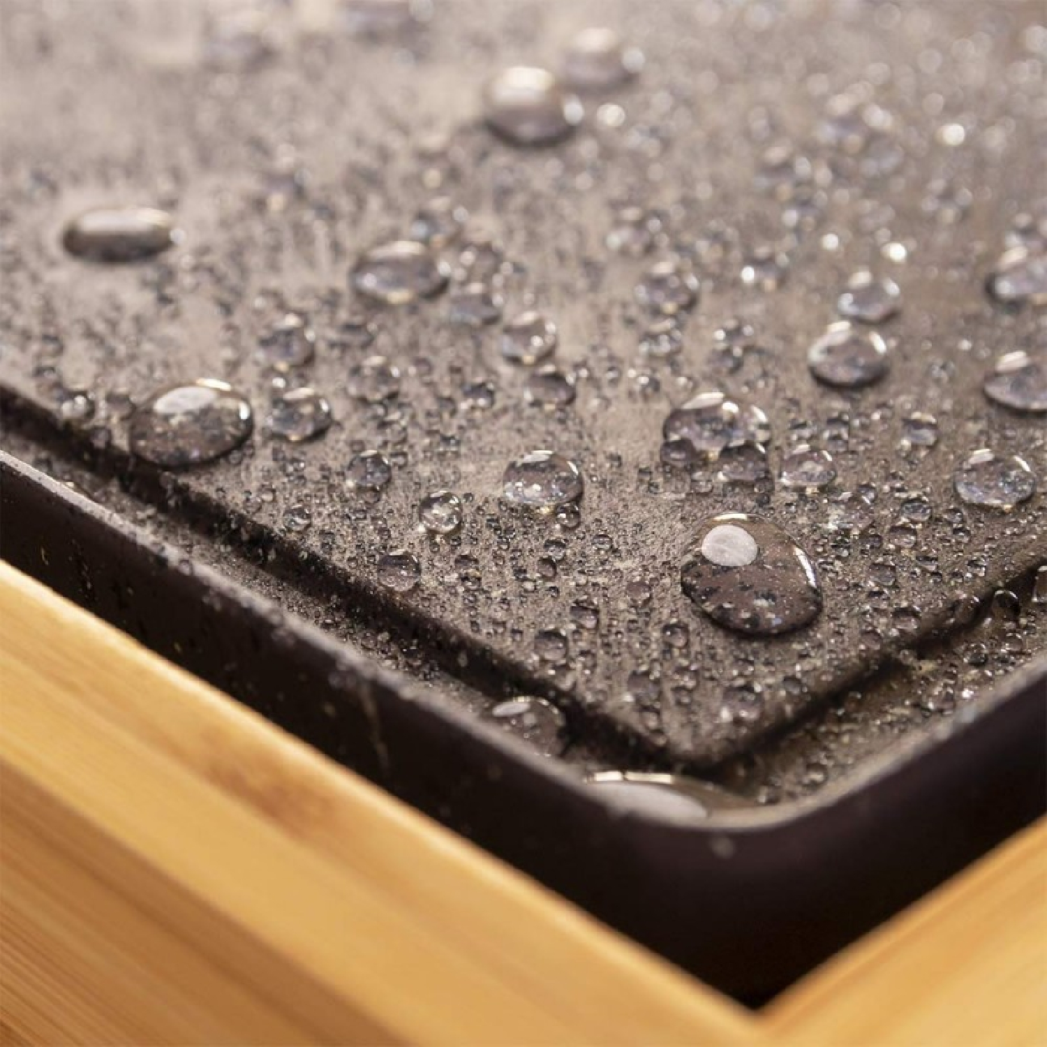 Close-up van een nat, donkergrijs oppervlak met veel waterdruppels. Het oppervlak wordt begrensd door een zwarte rand en een houten frame.