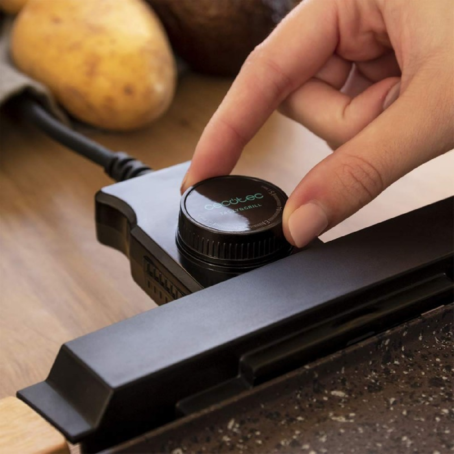 Een hand draait aan een zwarte knop op een grill. Op de achtergrond aardappelen. De knop is gelabeld 'Tasty & Grill'.