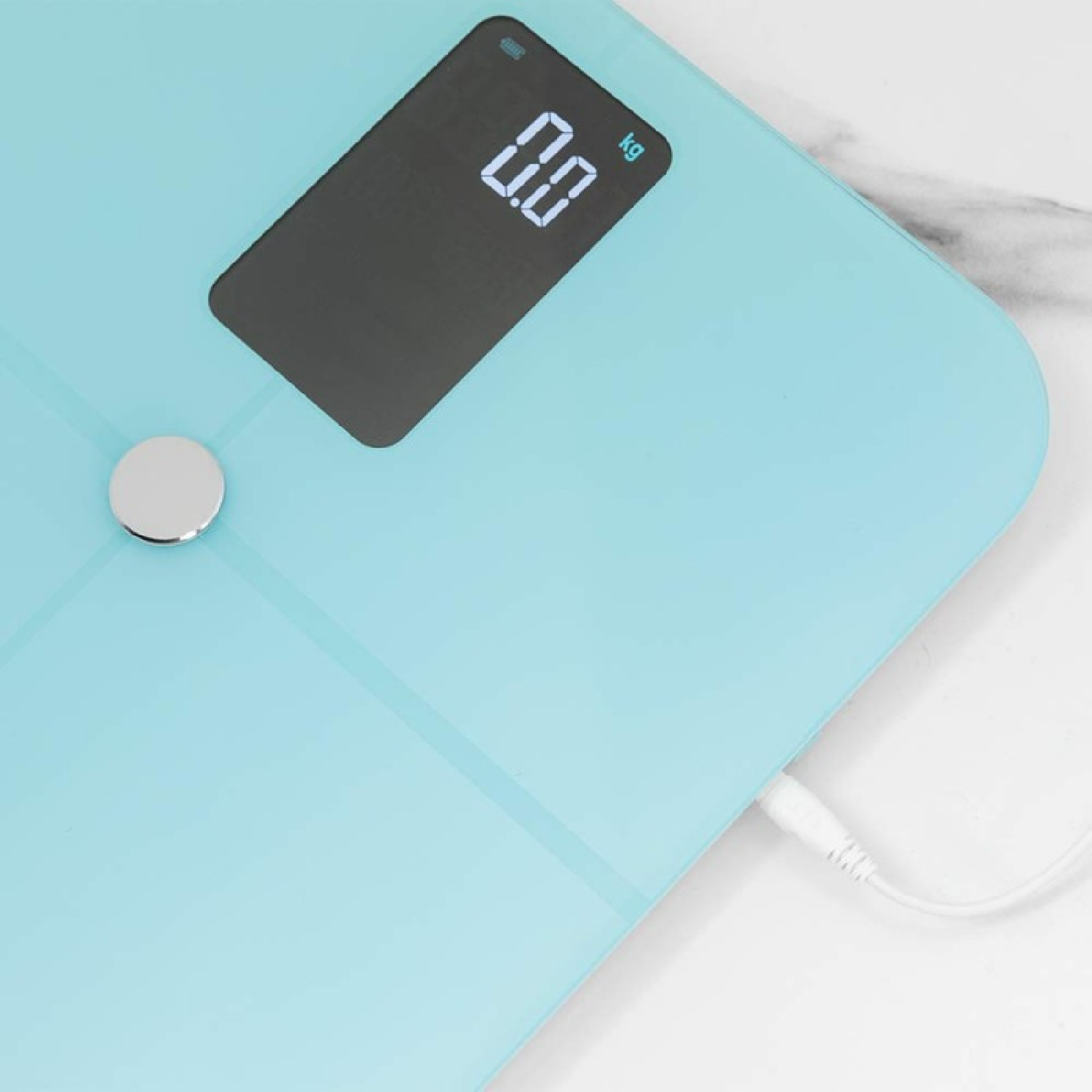 Una báscula de baño digital azul claro muestra 0.0 kg. Un botón plateado está a la izquierda. Un cable blanco está enchufado.