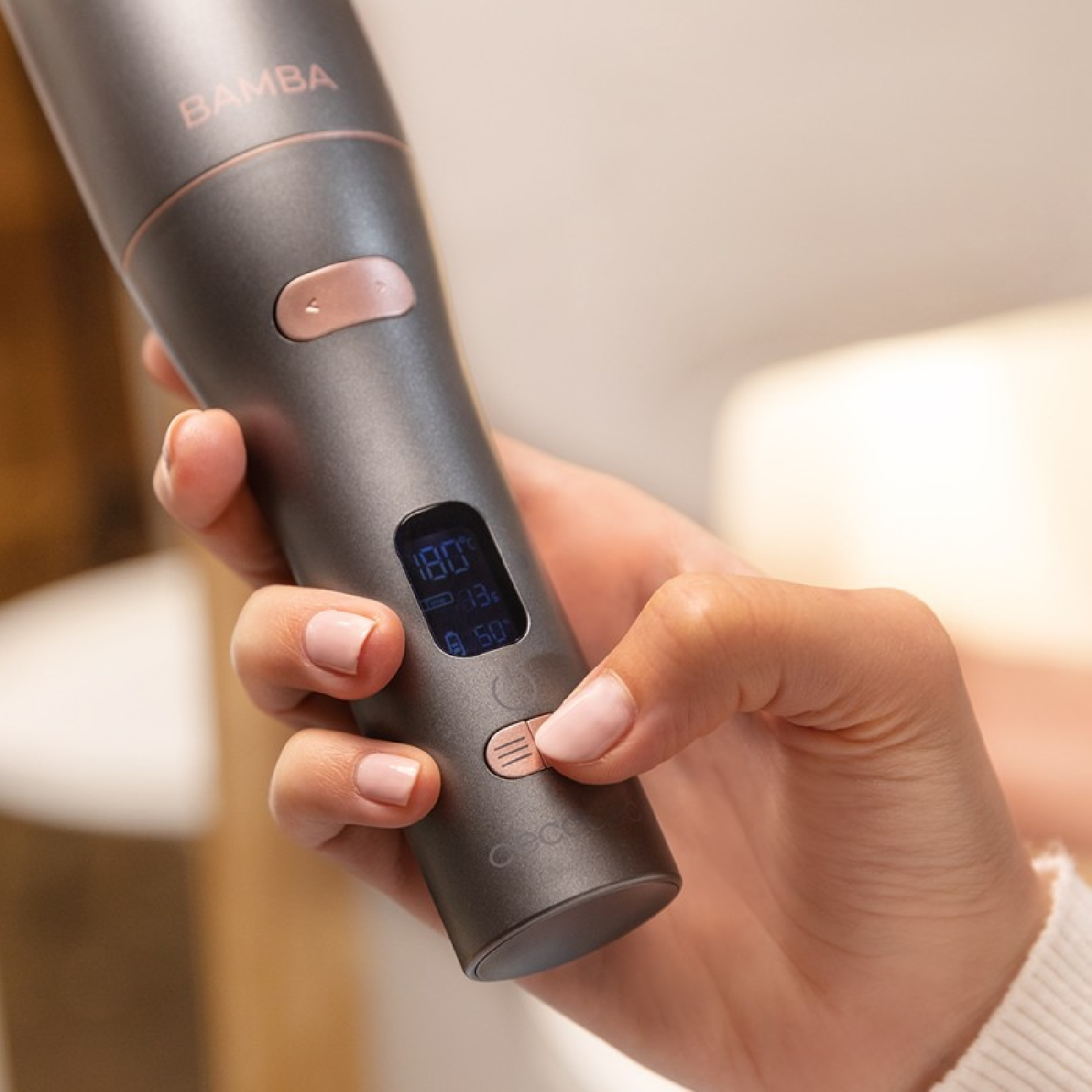 Eine Person hält einen Lockenstab, der die Temperatur anzeigt. Das Bügeleisen ist grau mit roségoldenen Akzenten. Ein digitales Display zeigt die Einstellungen.