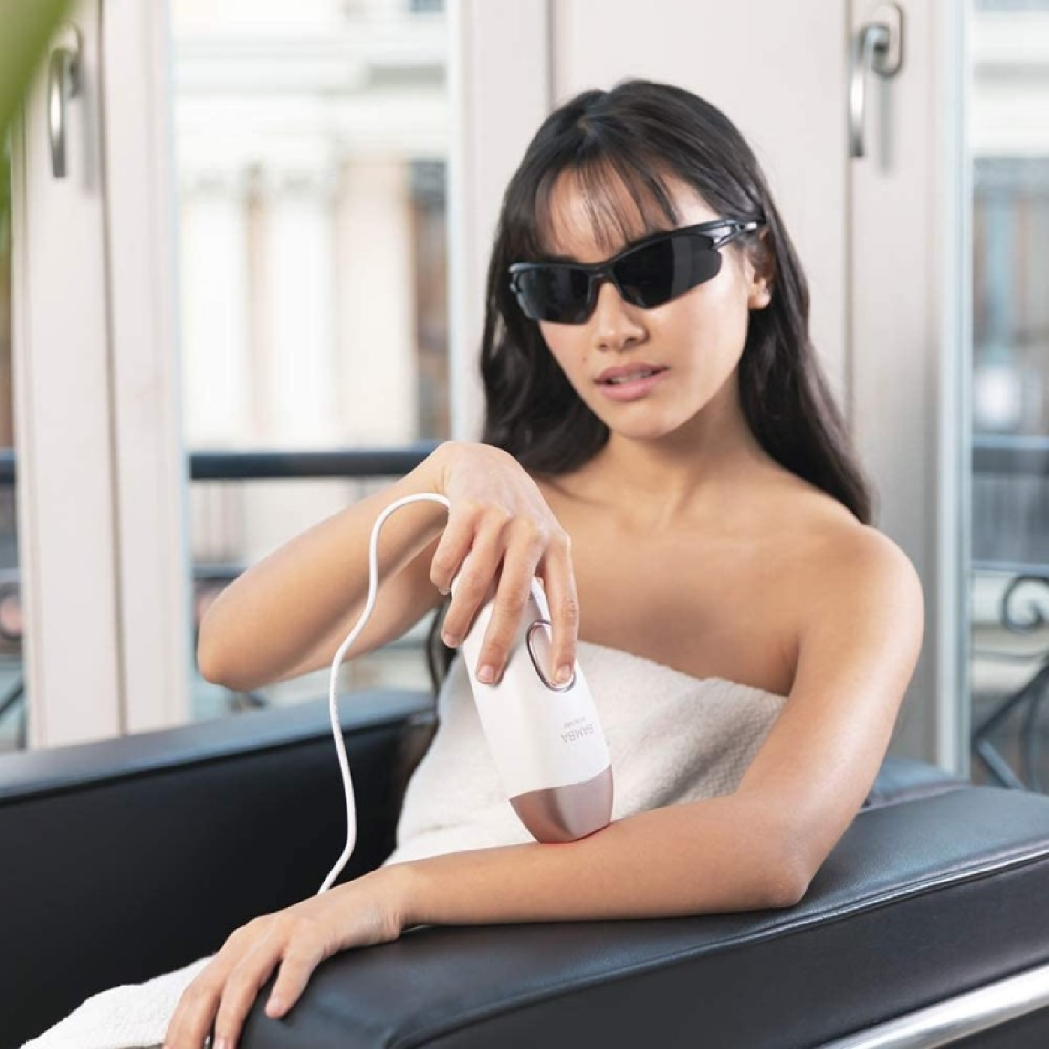 Kobieta w okularach przeciwsłonecznych używa urządzenia do usuwania włosów na ramieniu, siedząc na krześle. Tło przedstawia okna i balkon.