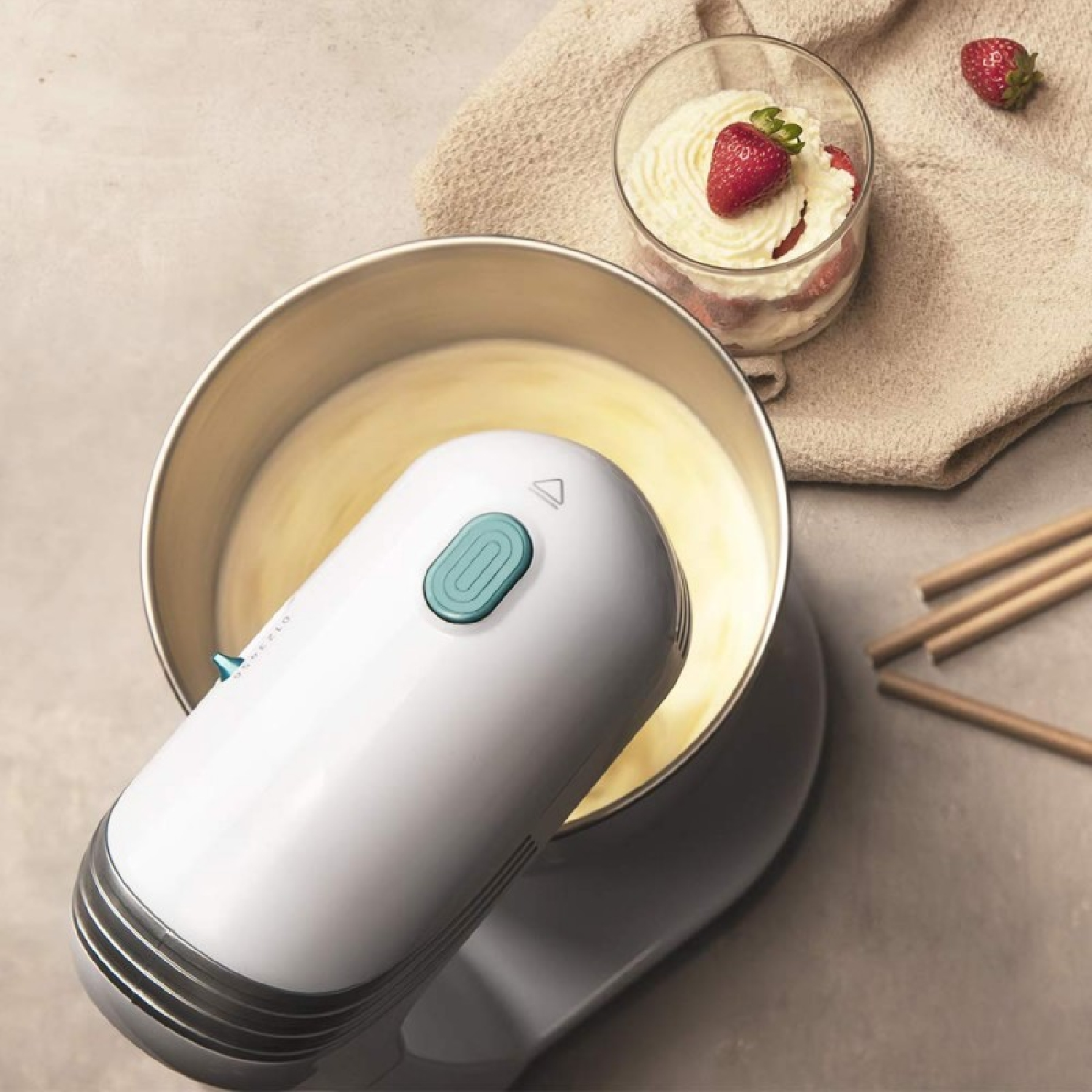 Un mixer bianco mescola l'impasto. Un dessert con fragole è a destra. Sono visibili anche bastoncini di legno.