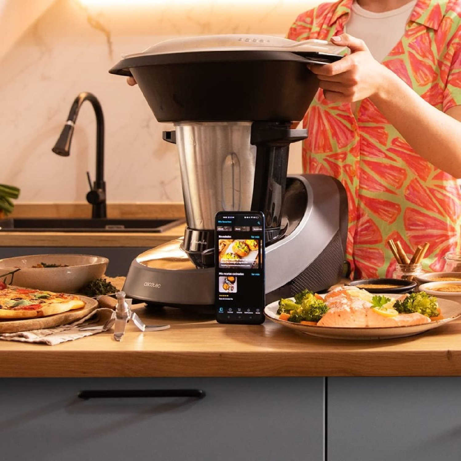 Una persona tiene un robot da cucina nero. Un telefono mostra ricette. Pizza, salmone e broccoli sul bancone.
