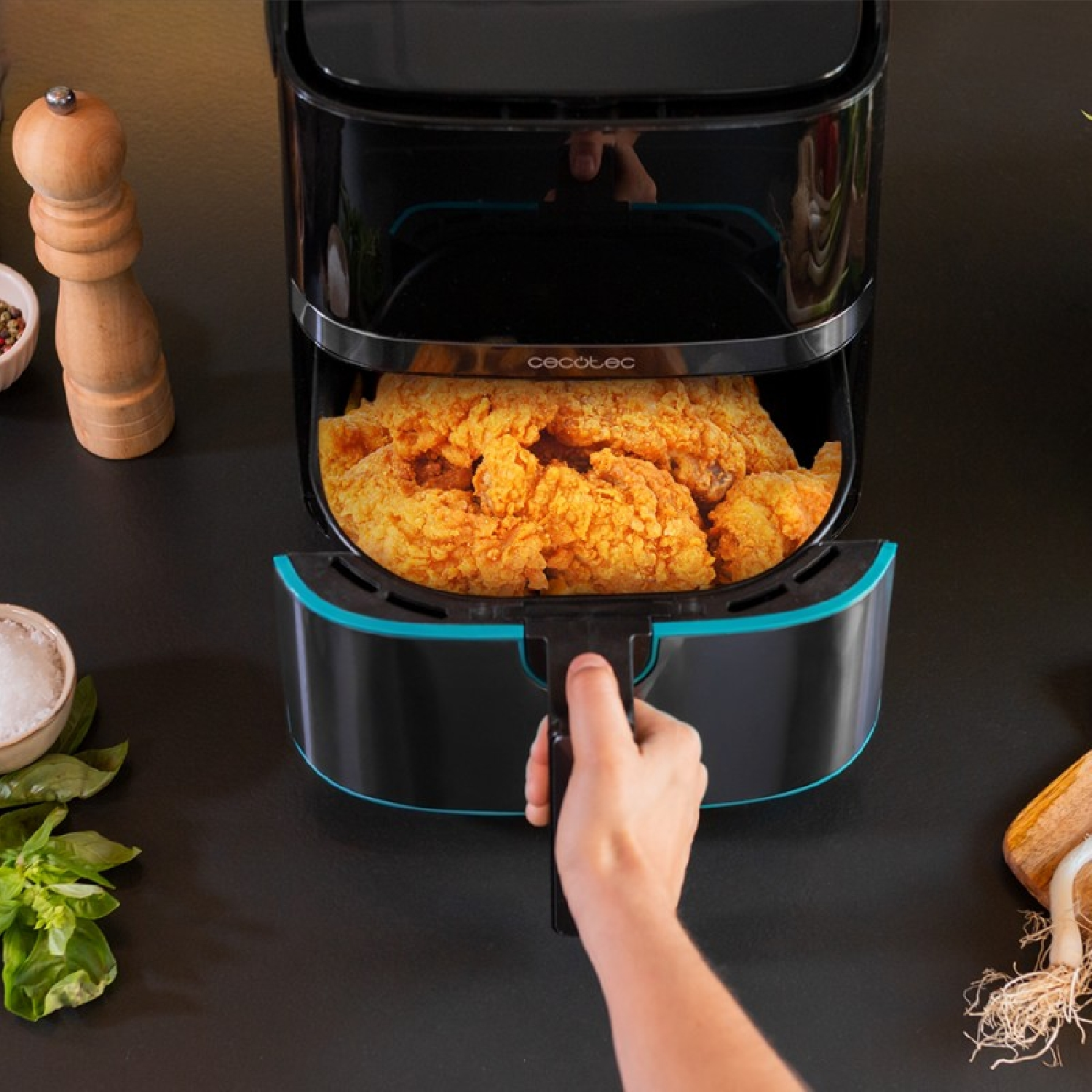 Una friggitrice ad aria piena di pollo fritto. Una mano tiene la maniglia. Sale, pepe ed erbe aromatiche accanto alla friggitrice.