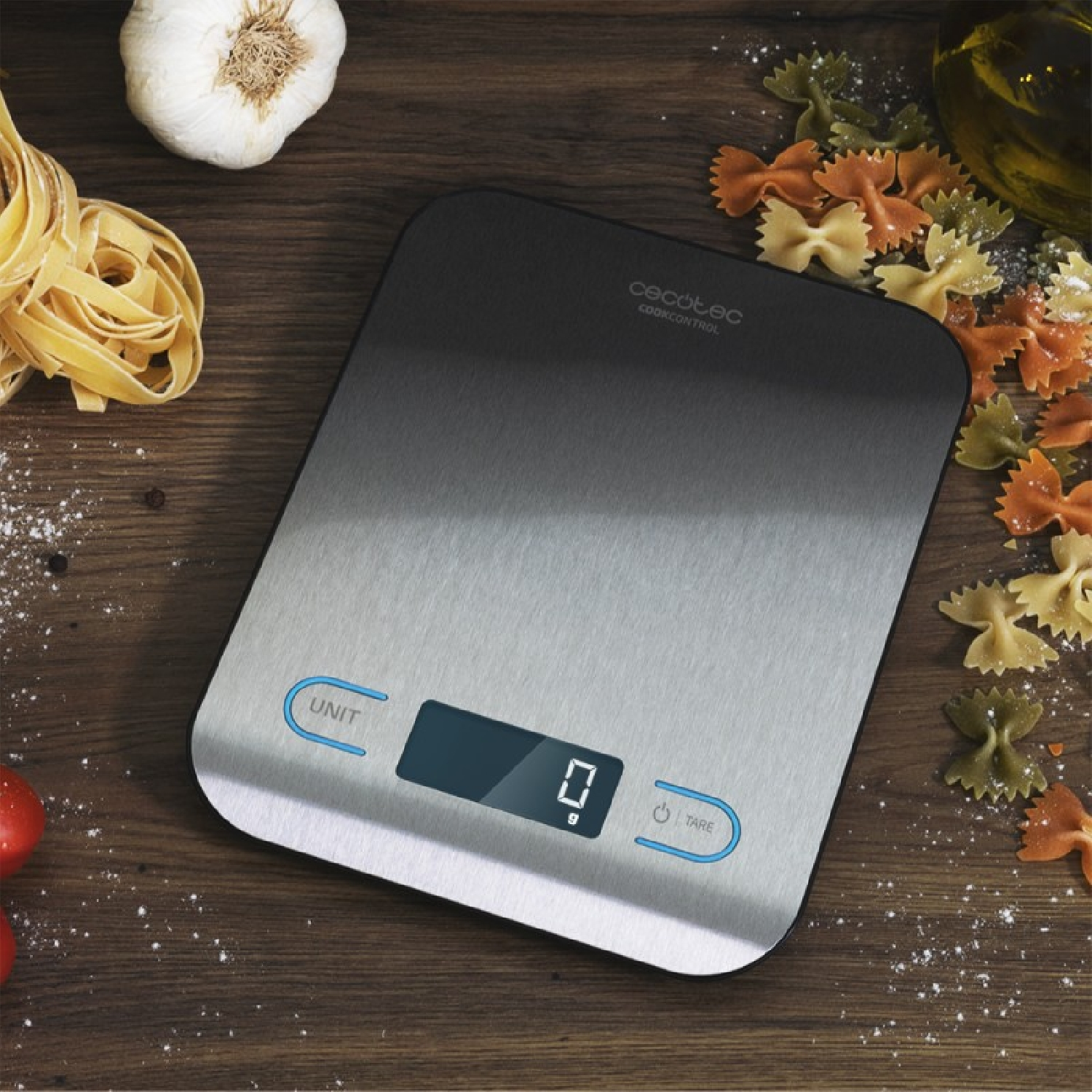 Una bilancia da cucina con cibo intorno. Aglio, pasta e olio sono visibili, su una superficie di legno.