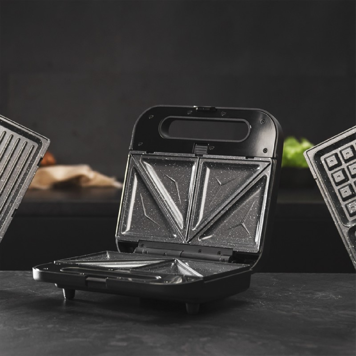 Sandwich maker nero aperto su una superficie scura. Due altre piastre su entrambi i lati. Lo sfondo è scuro.