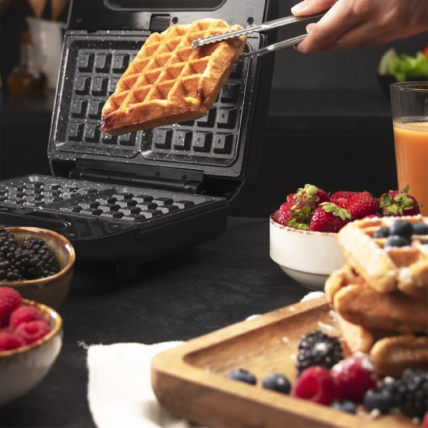 Un waffle viene sollevato da una macchina per waffle con delle pinze. Sono presenti anche bacche e succo, creando una scena di colazione.