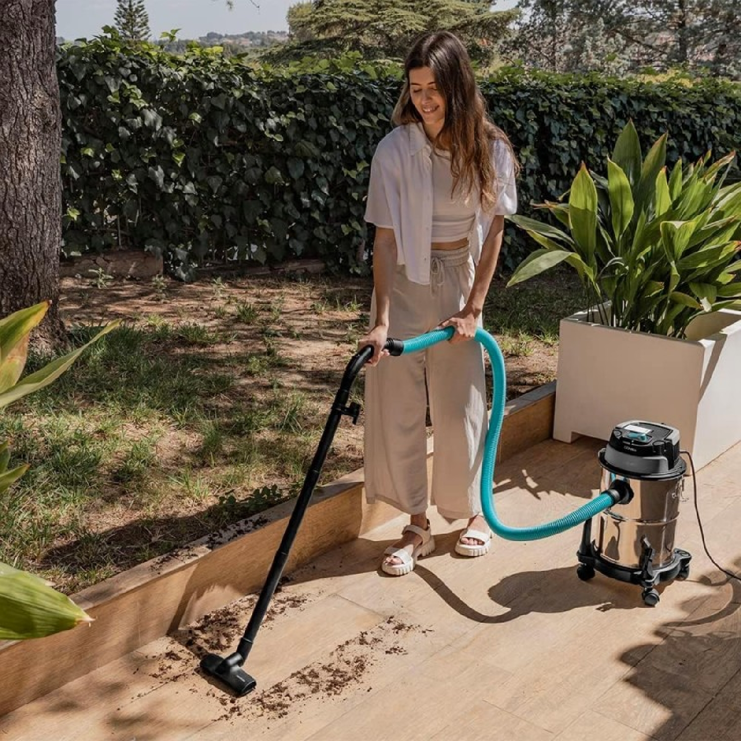 Una mujer aspira un patio. Lleva ropa blanca y beige. Se utiliza una aspiradora negra con una manguera azul.