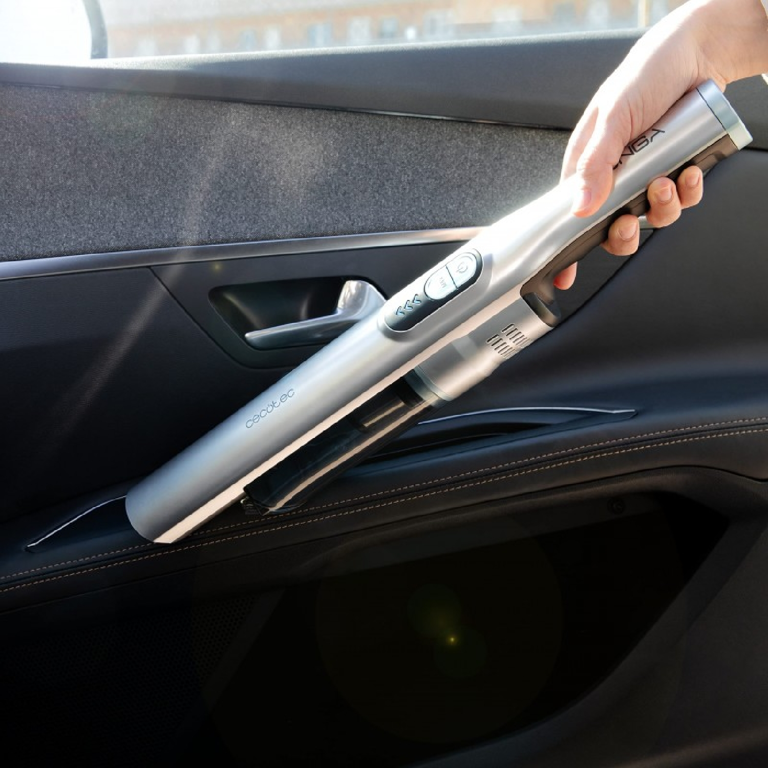 Una mano tiene un aspirapolvere Cecotec argentato all'interno di un'auto. La portiera è nera con tessuto grigio e cuciture marroni.