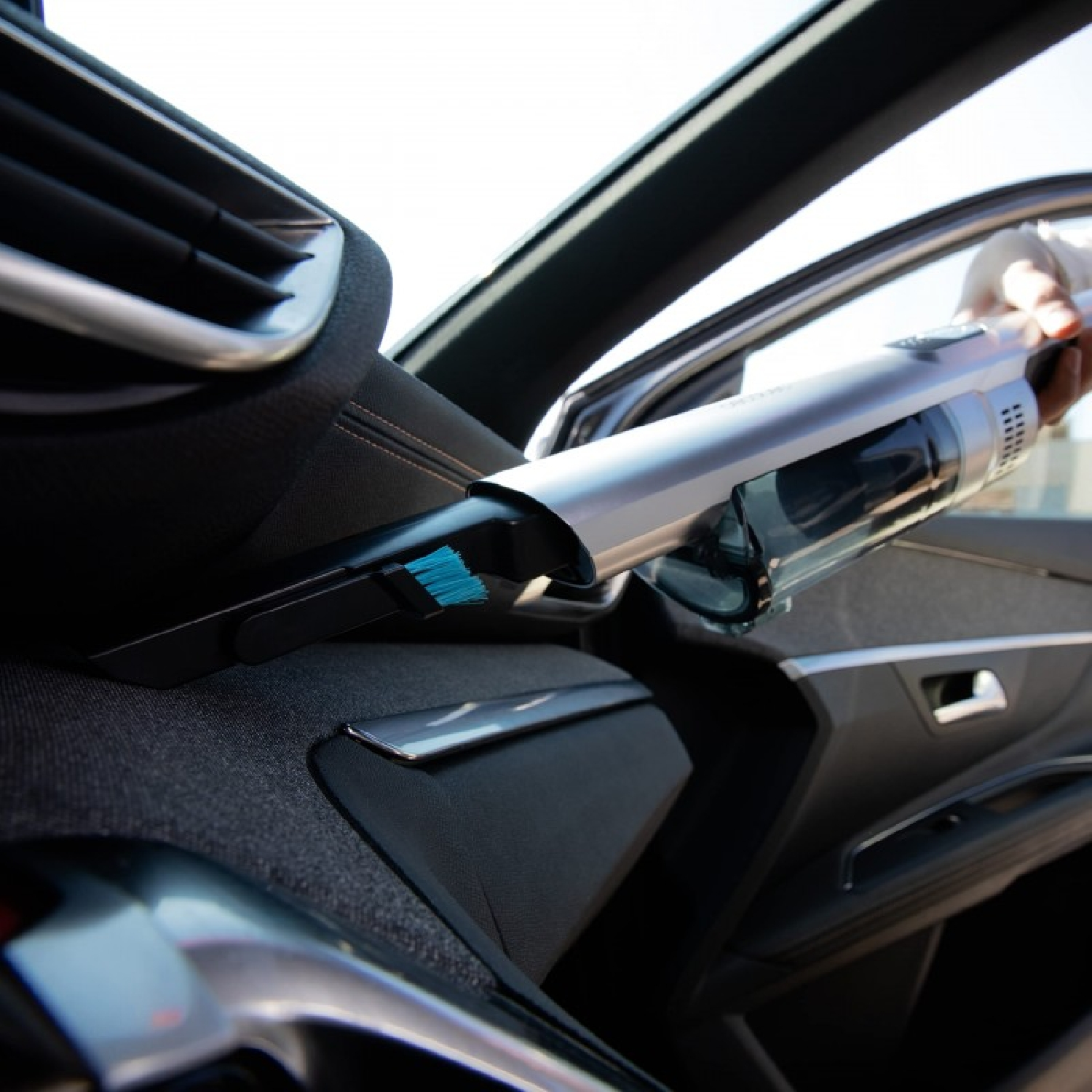 Una persona aspira l'interno di un'auto con un aspirapolvere portatile. L'attenzione è sull'interno dell'auto e sull'aspirapolvere.