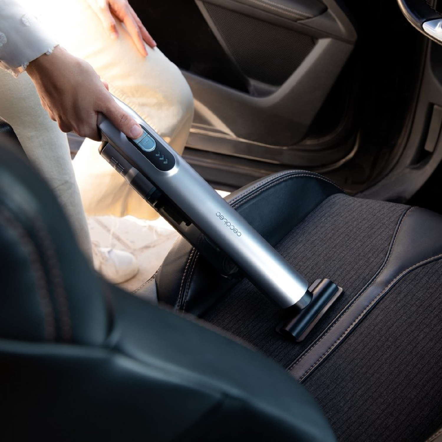 Una persona aspira un sedile dell'auto con un aspirapolvere portatile argentato. Il sedile è nero e l'interno dell'auto è visibile.