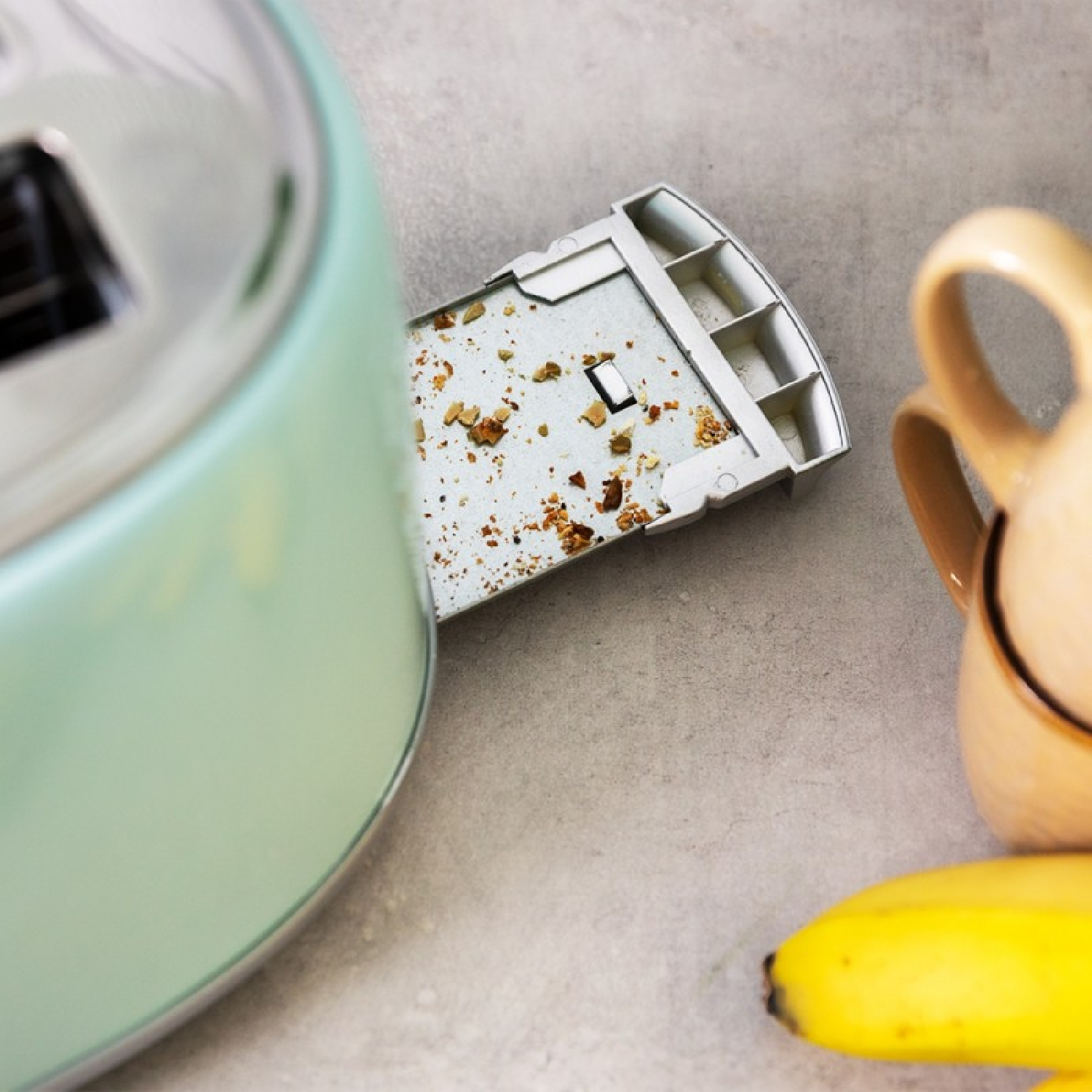 Ein mintgrüner und verchromter Toaster mit Krümelschublade, ein beiger Becher und eine gelbe Banane auf grauem Untergrund.
