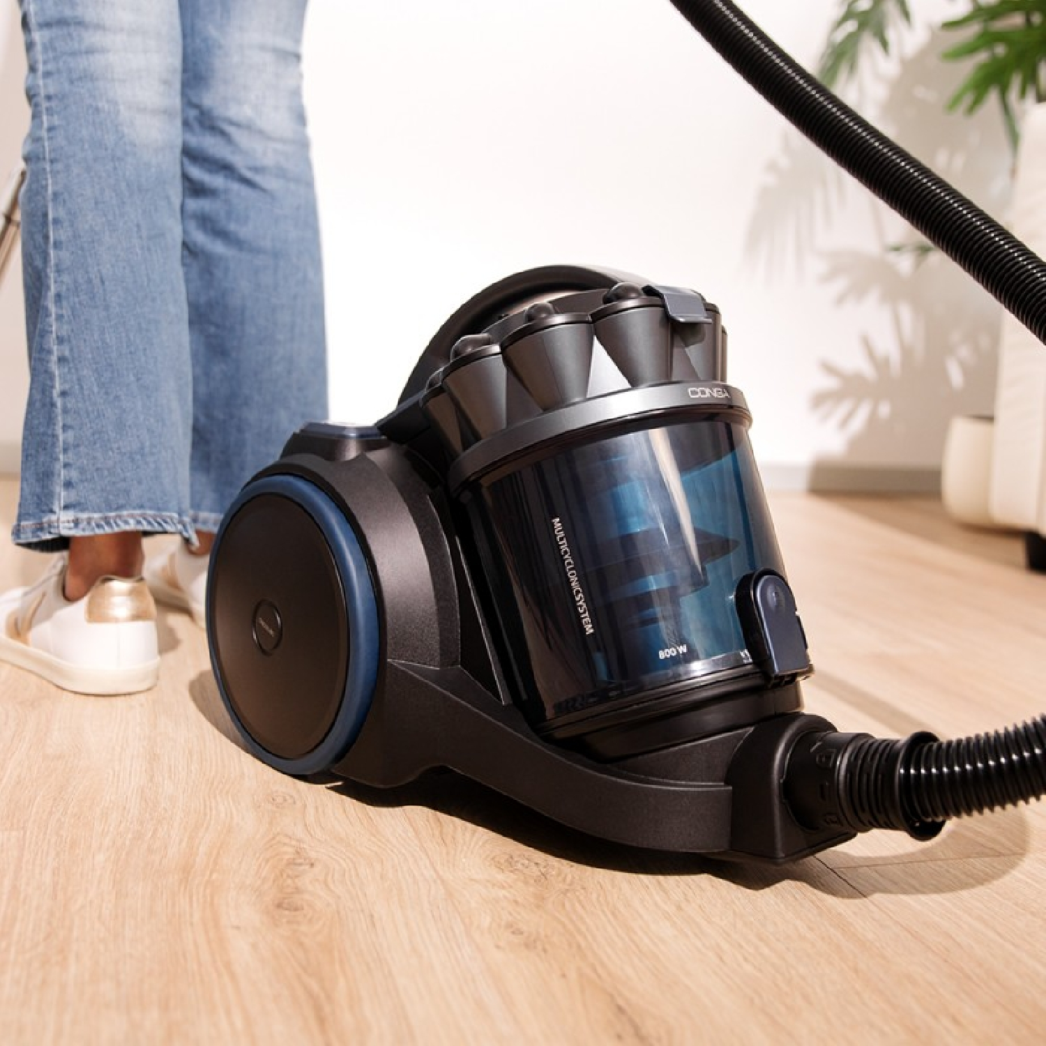 Een persoon stofzuigt een houten vloer met een zwart-blauwe stofzuiger. De persoon draagt een blauwe spijkerbroek en witte schoenen.