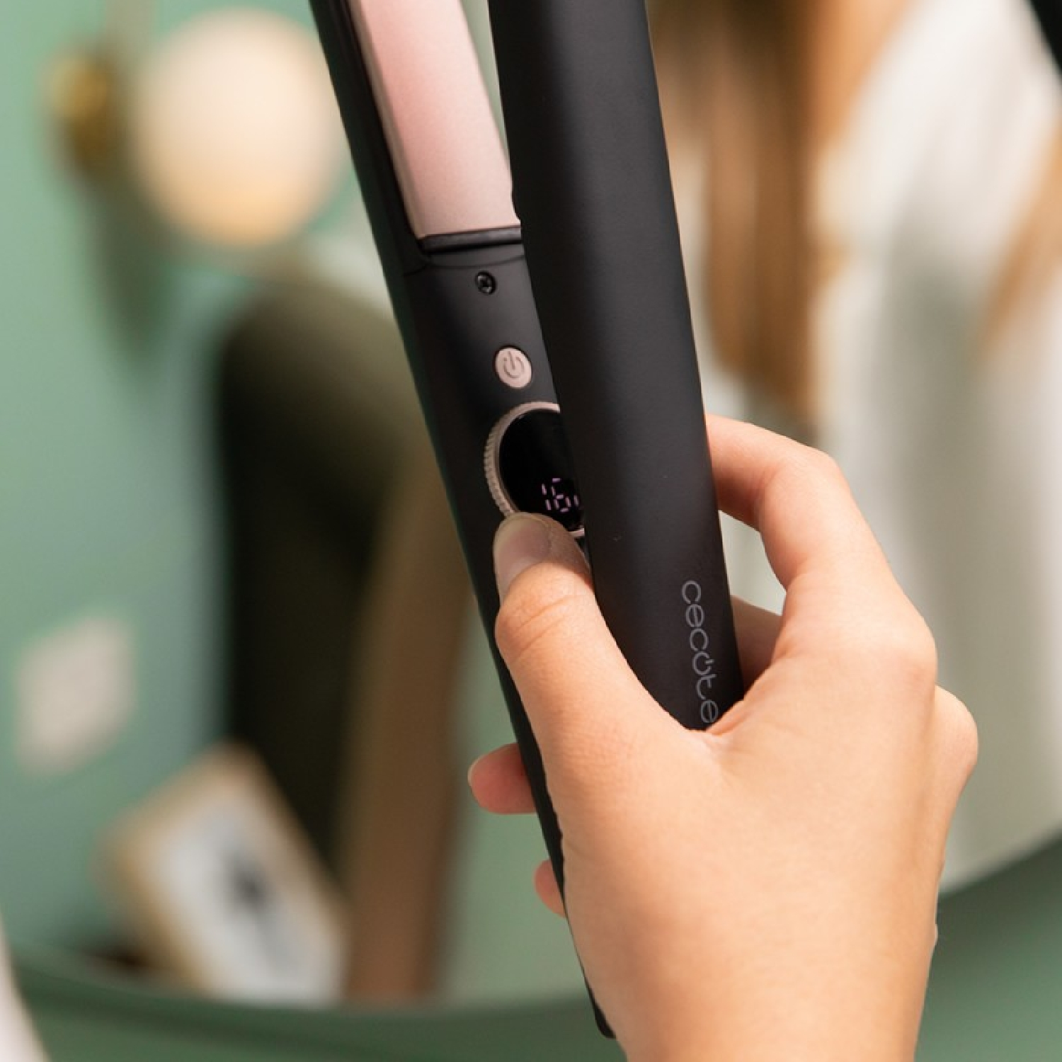 Una mano tiene in mano una piastra per capelli nera con display digitale che mostra 160. Lo sfondo è uno specchio e un muro sfocati.