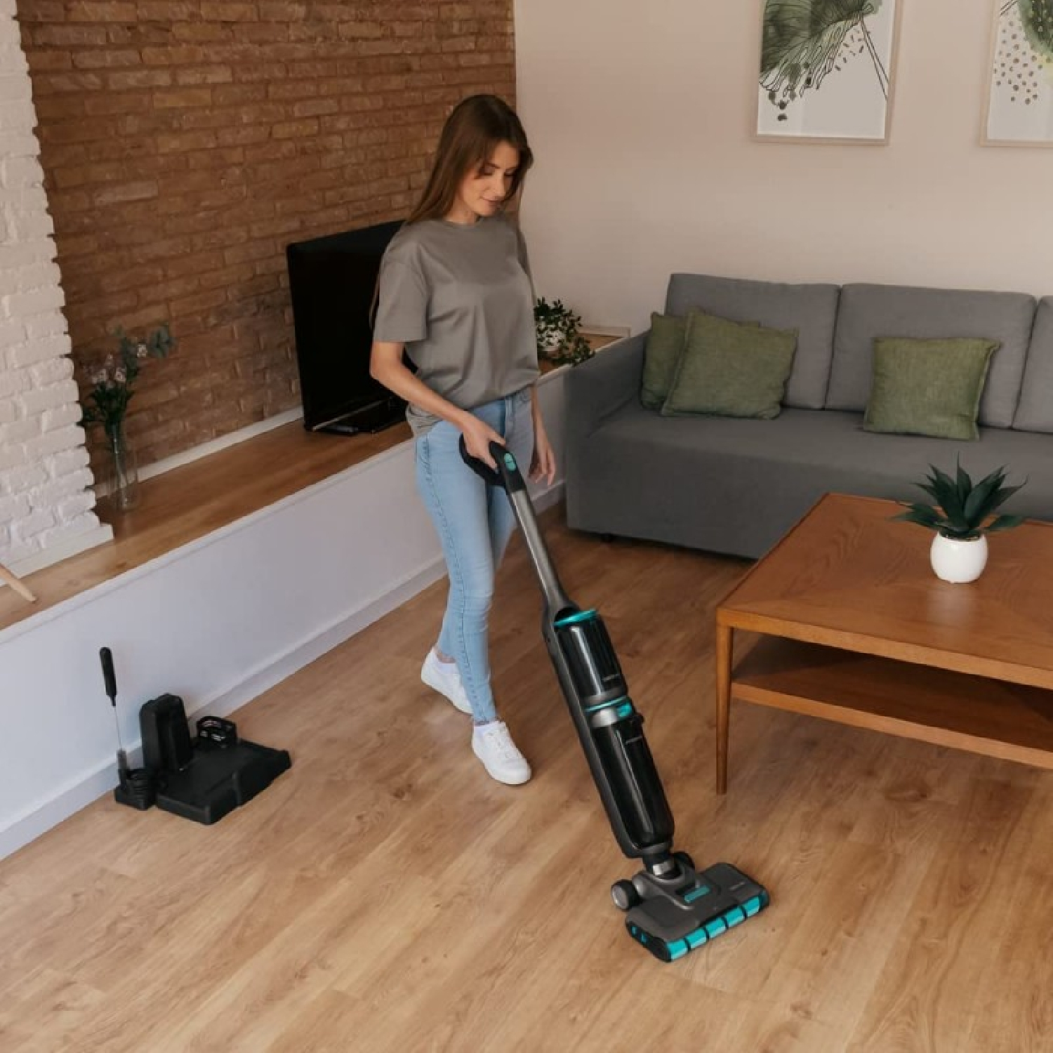Una mujer aspira un suelo de madera. Hay un sofá, una mesa y una pared de ladrillo al fondo.