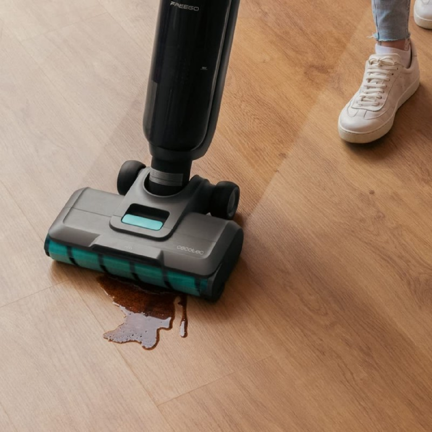 Un limpiador de pisos está limpiando un derrame de líquido marrón en un piso de madera. Una persona con zapatillas blancas está cerca.