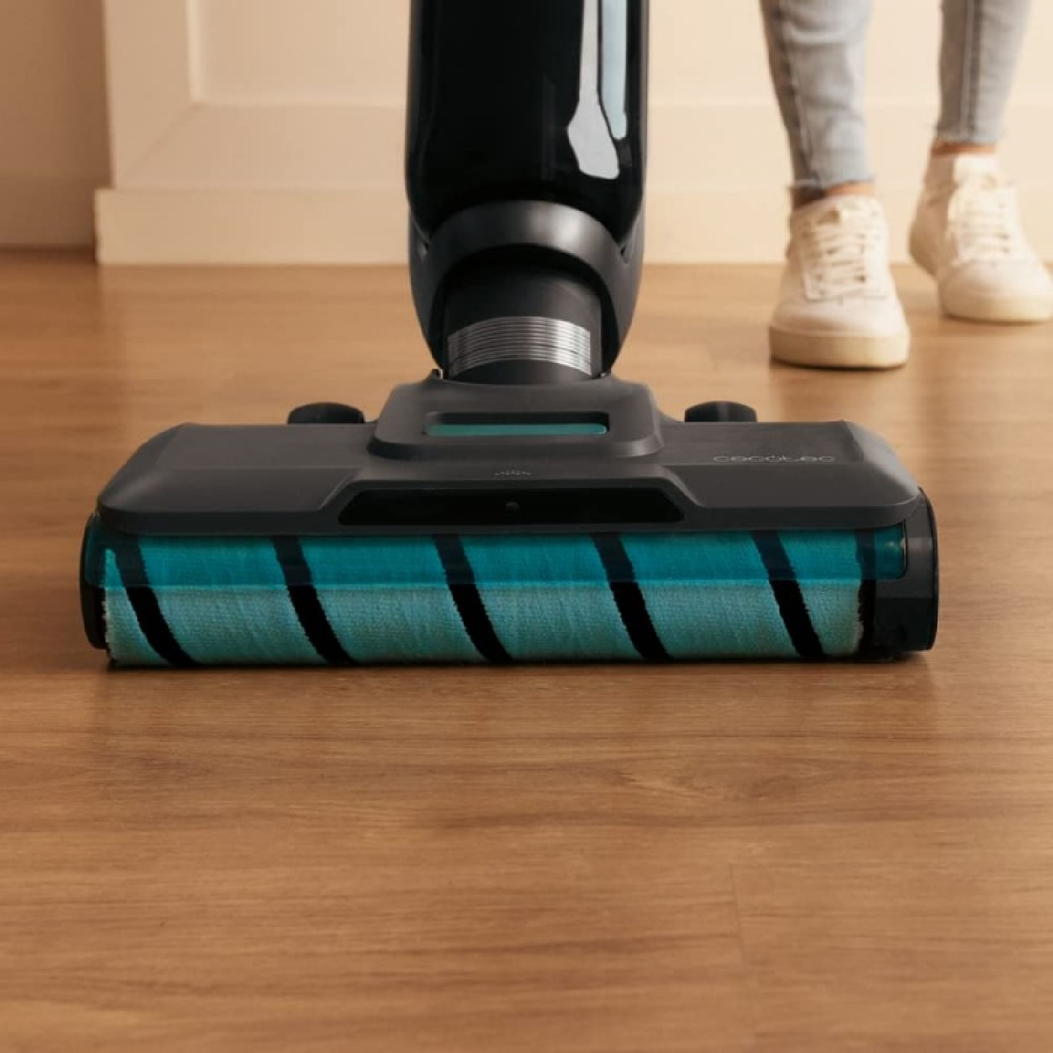 Una aspiradora con un rodillo turquesa y negro está sobre un piso de madera. Una persona en jeans y zapatillas blancas está detrás.