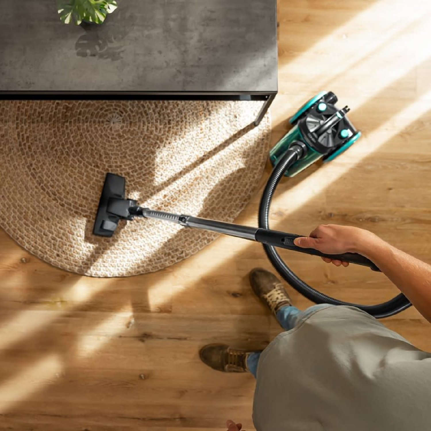 Osoba odkurza pokój. Czarna głowica odkurzacza czyści dywan. Odkurzacz jest zielono-czarny. Podłoga jest drewniana.
