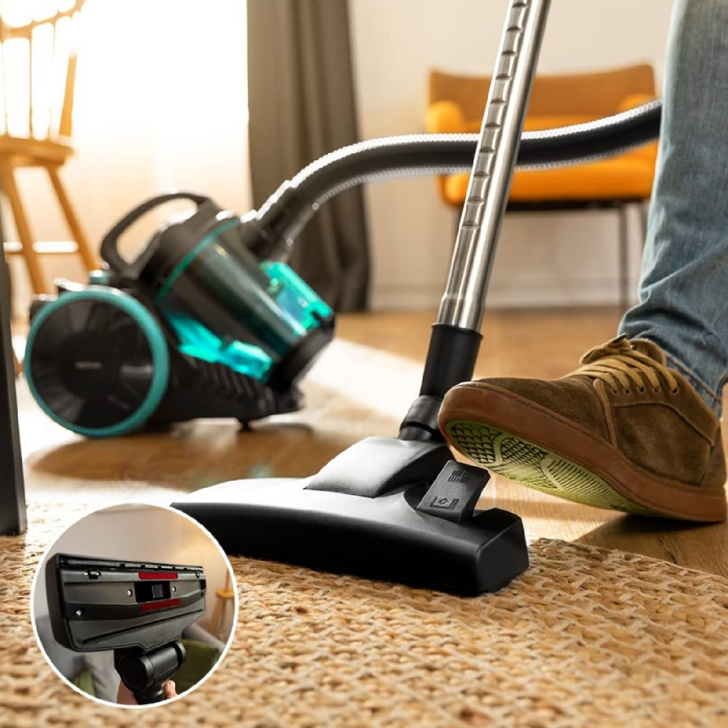 Osoba odkurza dywan czarnym i turkusowym odkurzaczem. Stopa osoby znajduje się na głowicy odkurzacza.