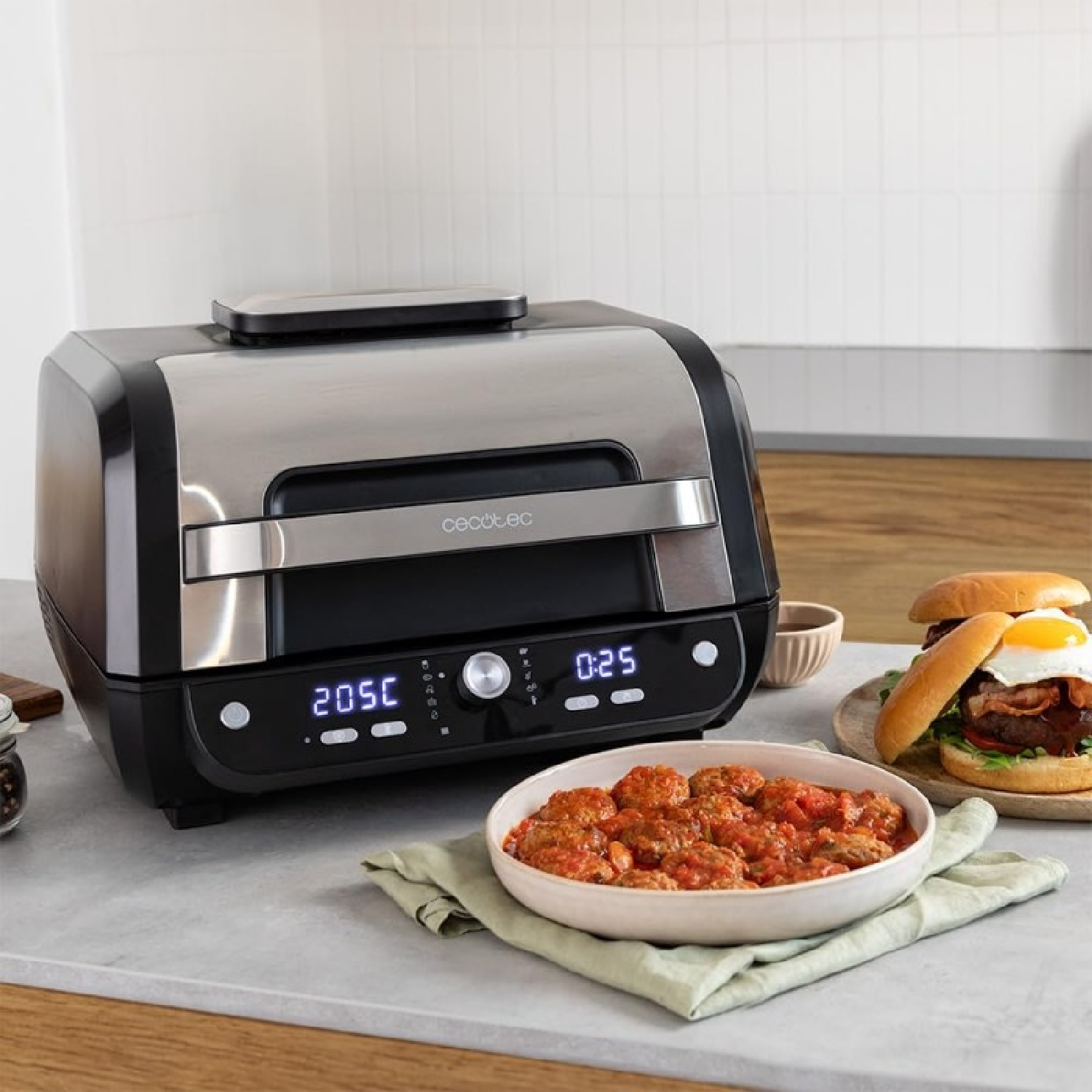 Una parrilla eléctrica con albóndigas y una hamburguesa en la mesa. La parrilla es negra y plateada. La hamburguesa tiene un huevo frito.