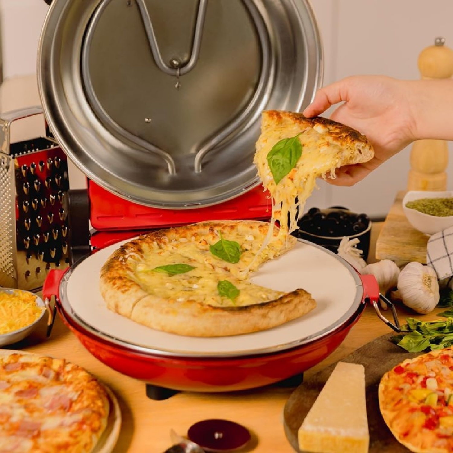 Una mano tiene una fetta di pizza con formaggio fuso. Altre pizze, ingredienti e un forno per pizza su un tavolo di legno.
