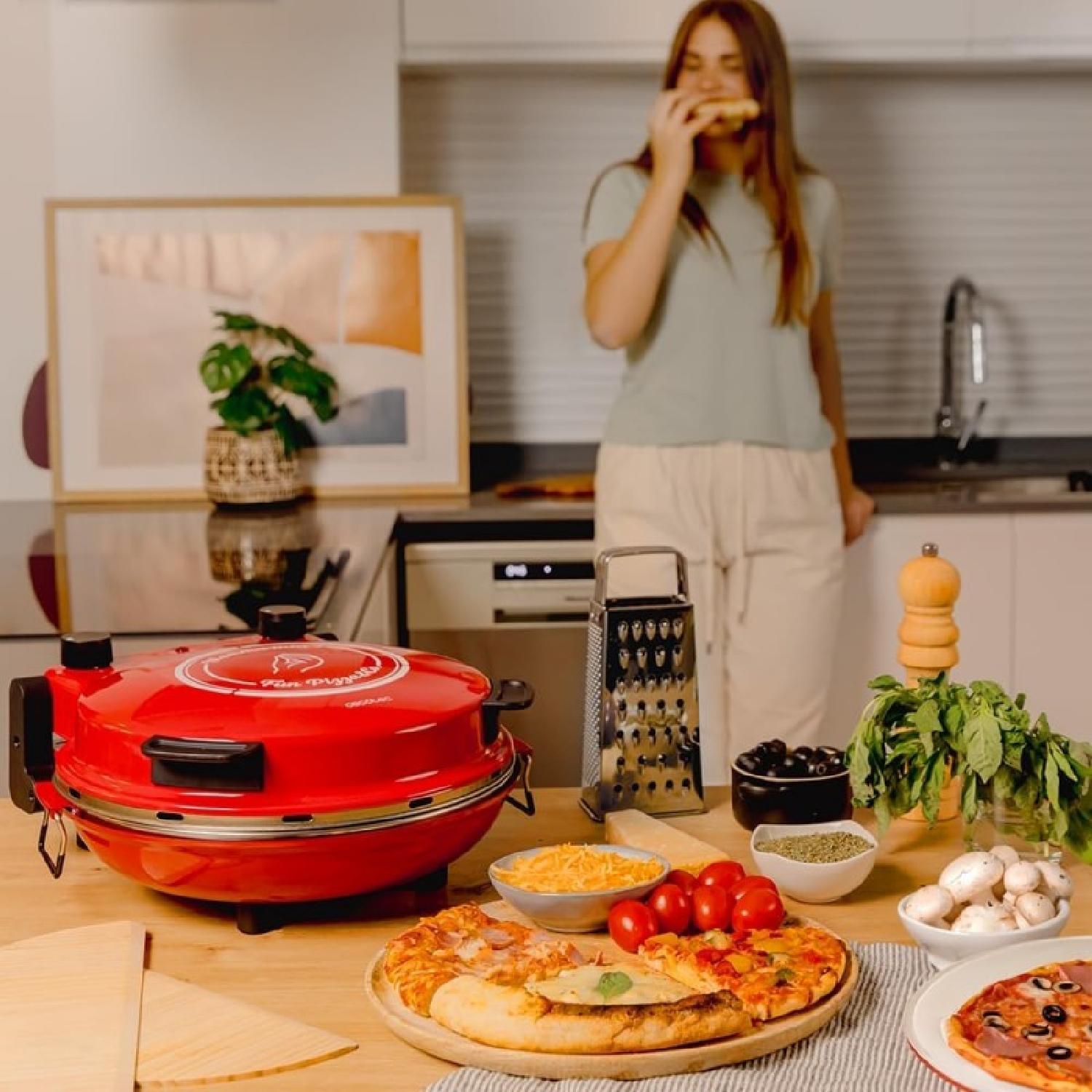 Una donna mangia pizza vicino a un forno per pizza rosso. Pizza, ingredienti e una grattugia sono sul tavolo. Un quadro è appeso al muro.