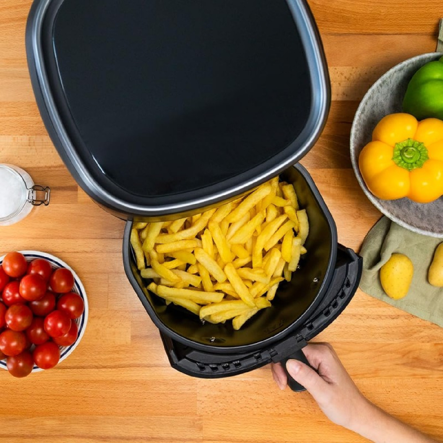 Una freidora de aire con papas fritas, tomates, pimientos y patatas sobre madera. Una mano sostiene la freidora.