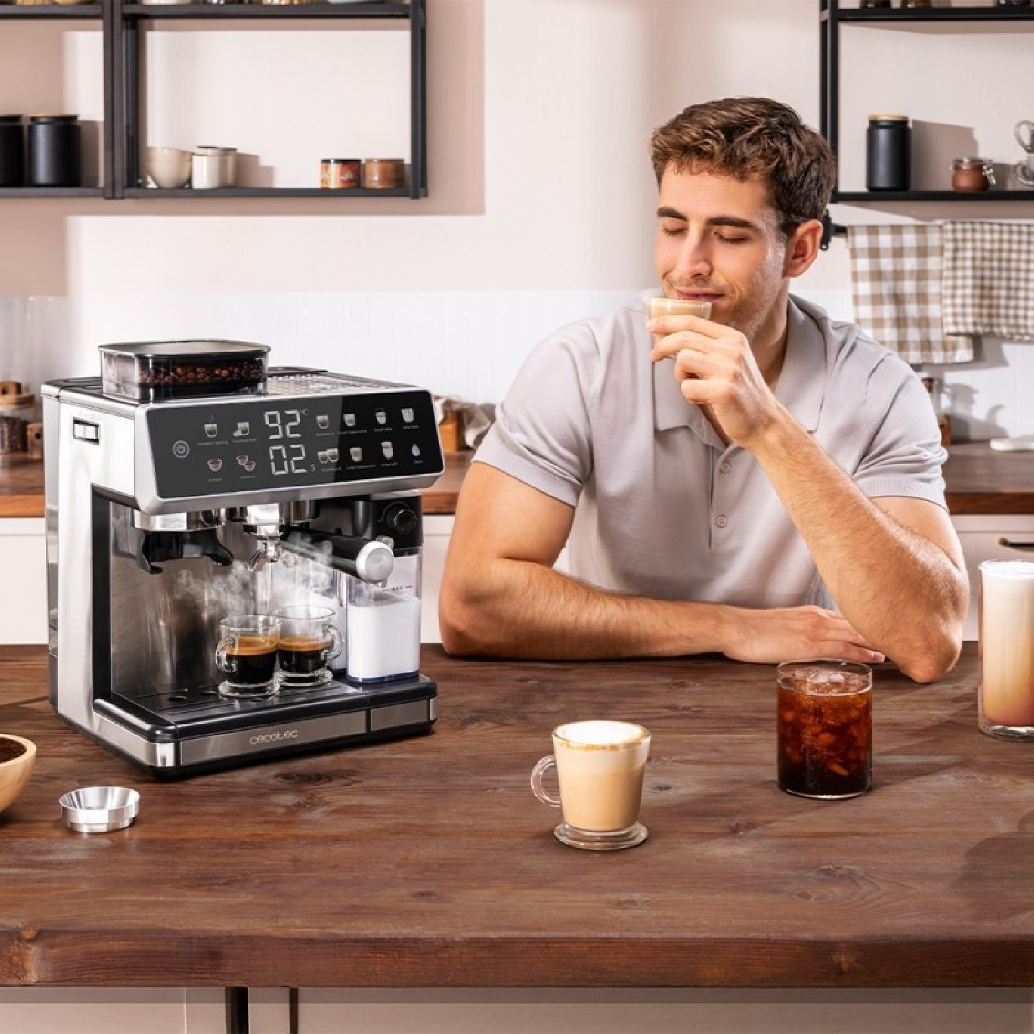 Uomo che assaggia il caffè, macchina per caffè espresso, varie bevande al caffè su un tavolo di legno. Ambiente cucina. Uomo in camicia grigia.