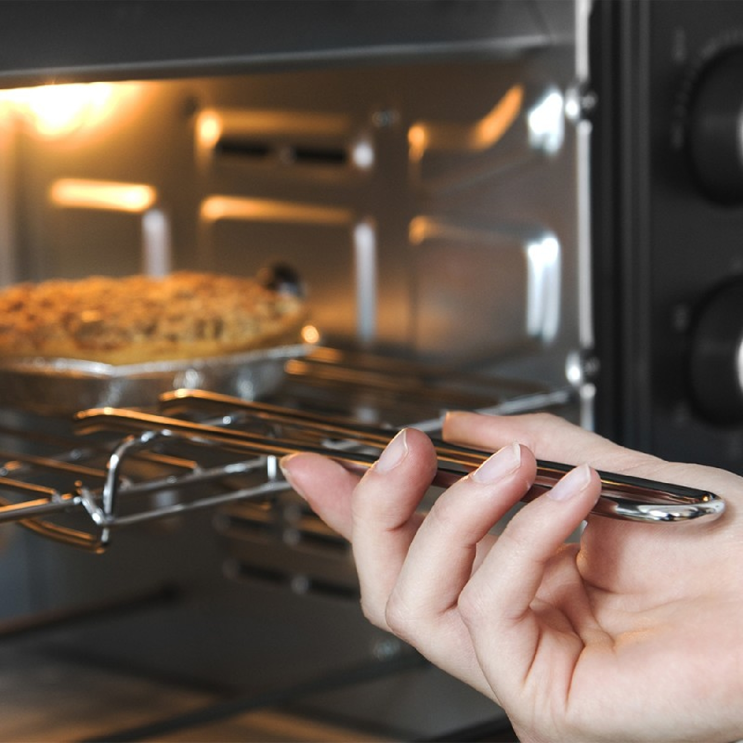 Una mano tira fuori una griglia metallica dal forno. All'interno, un piatto di cibo cuoce sotto una luce calda.