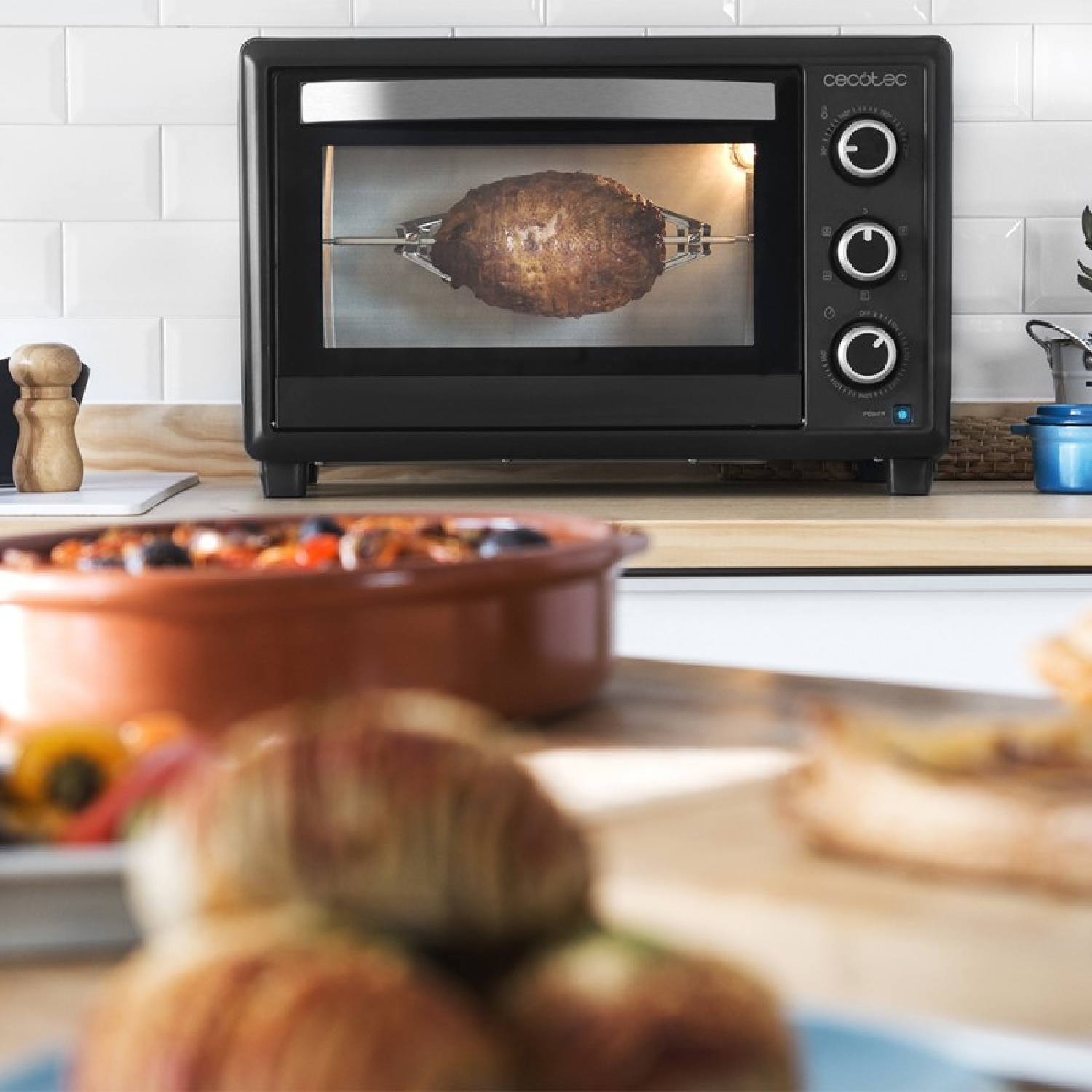 Un forno nero con un pollo arrosto all'interno. Altri cibi e utensili da cucina sono in primo e secondo piano.