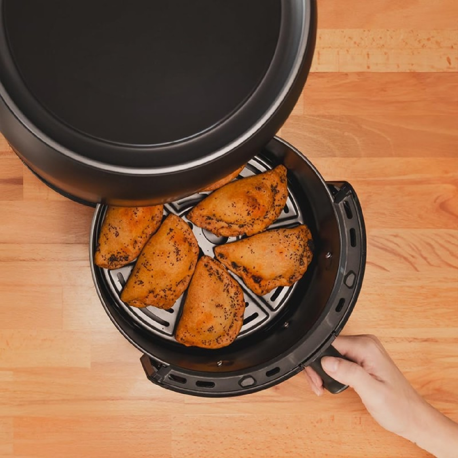 Una freidora de aire con tapa abierta, revelando pasteles dorados. Una mano sostiene la freidora. Fondo de mesa de madera.
